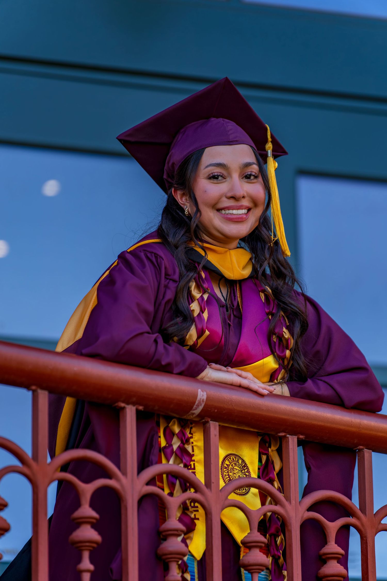 Graduate in purple commencement attire smiles while leaning on ornate metal railing.