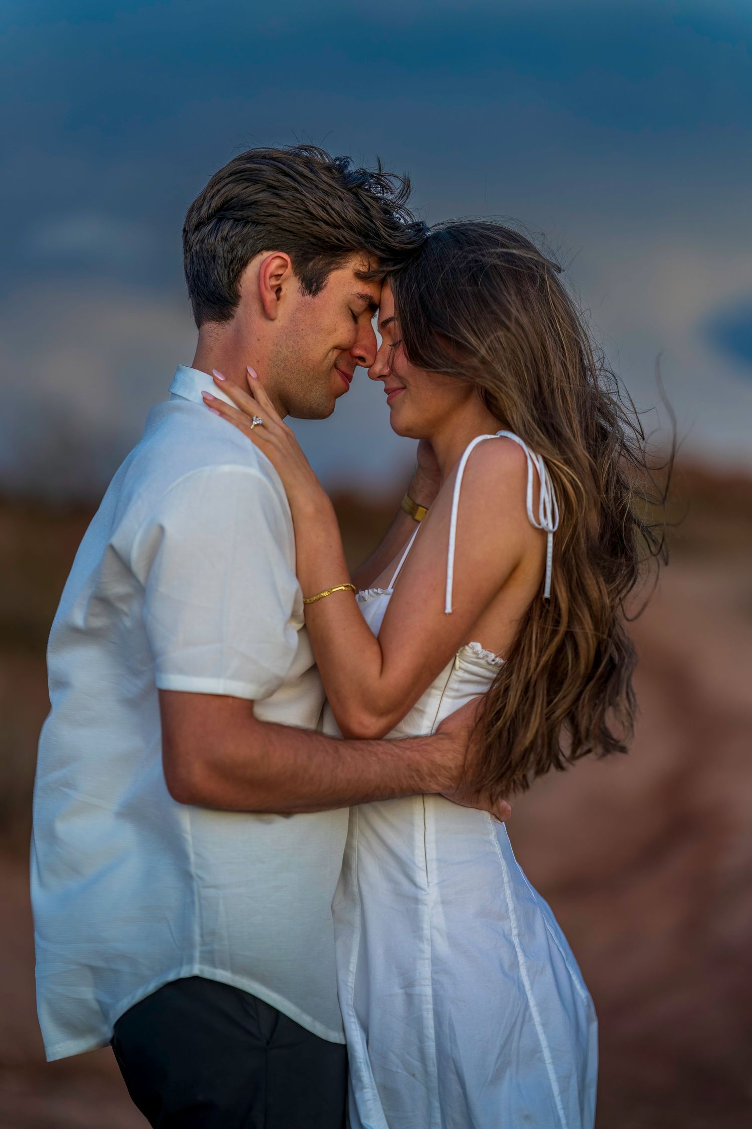 Couple in white outfits sharing a tender moment against a dramatic sky backdrop.