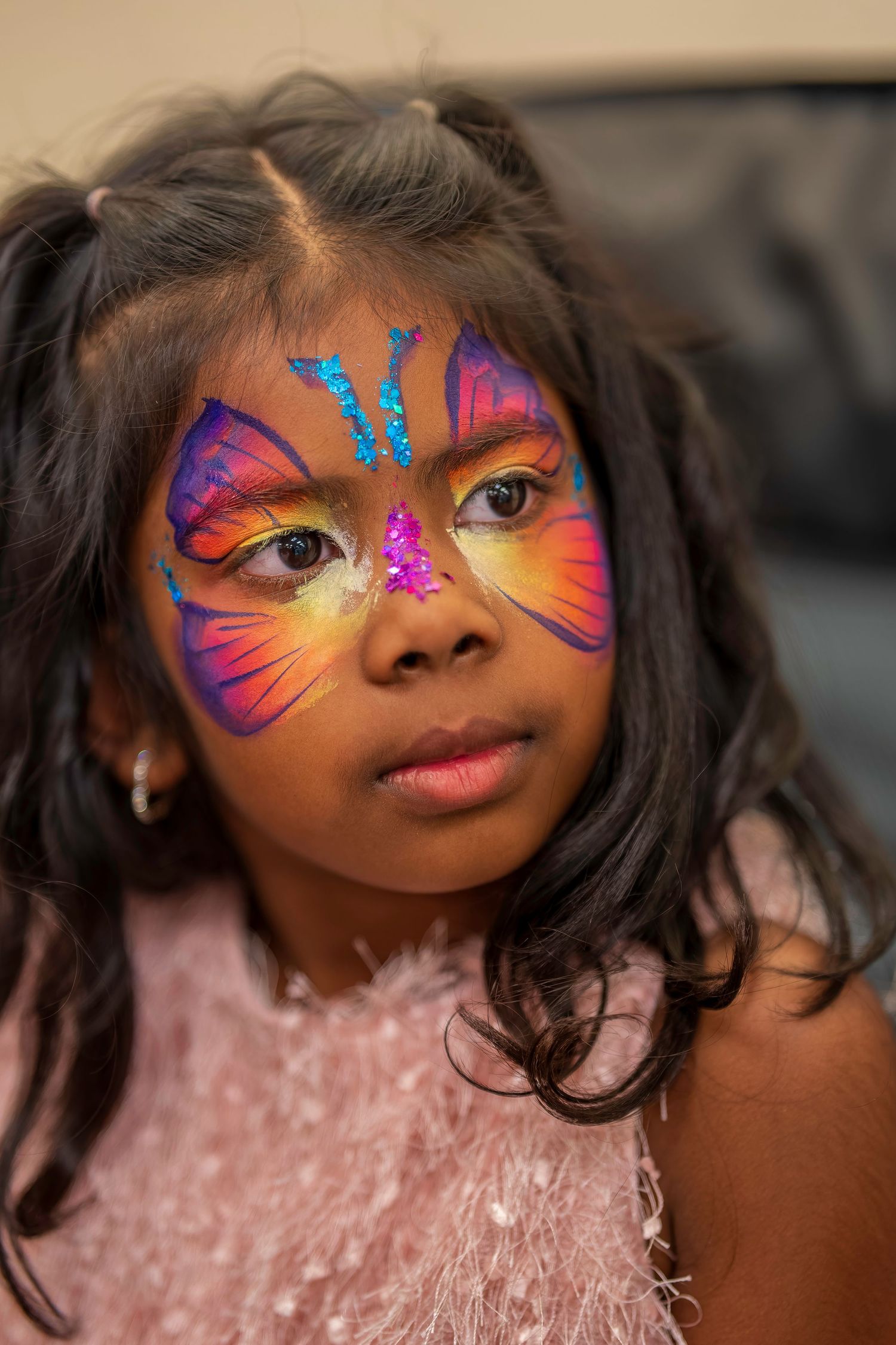Young girl with vibrant butterfly face paint in pink, orange, and purple with blue glitter accents.