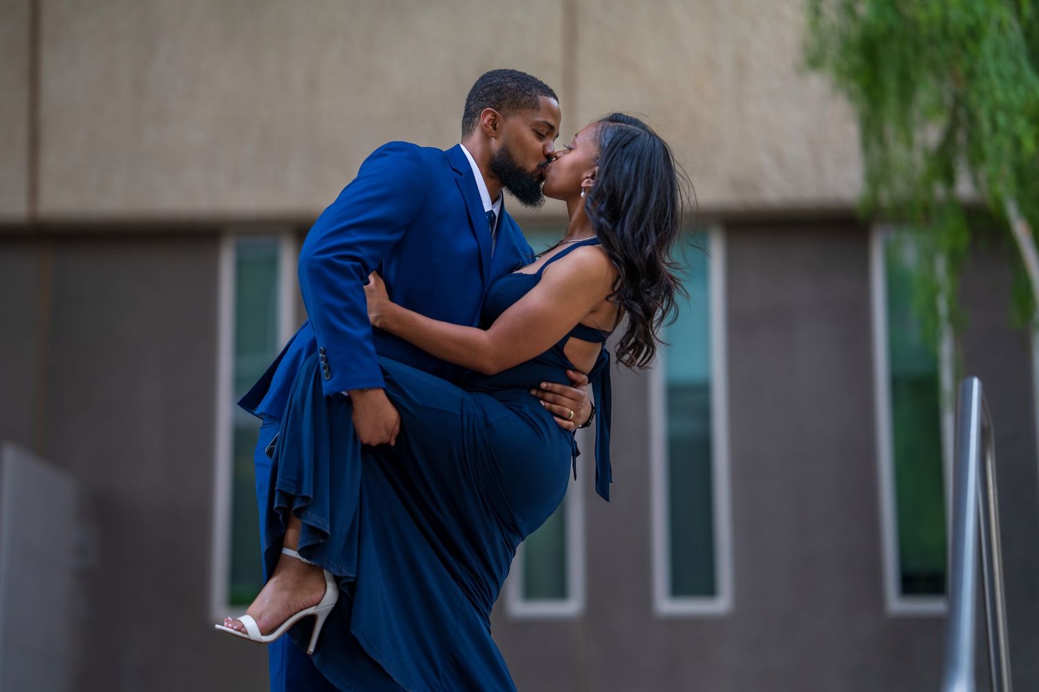 Couple in matching blue formal attire sharing a kiss, man lifting woman outdoors at dusk.
