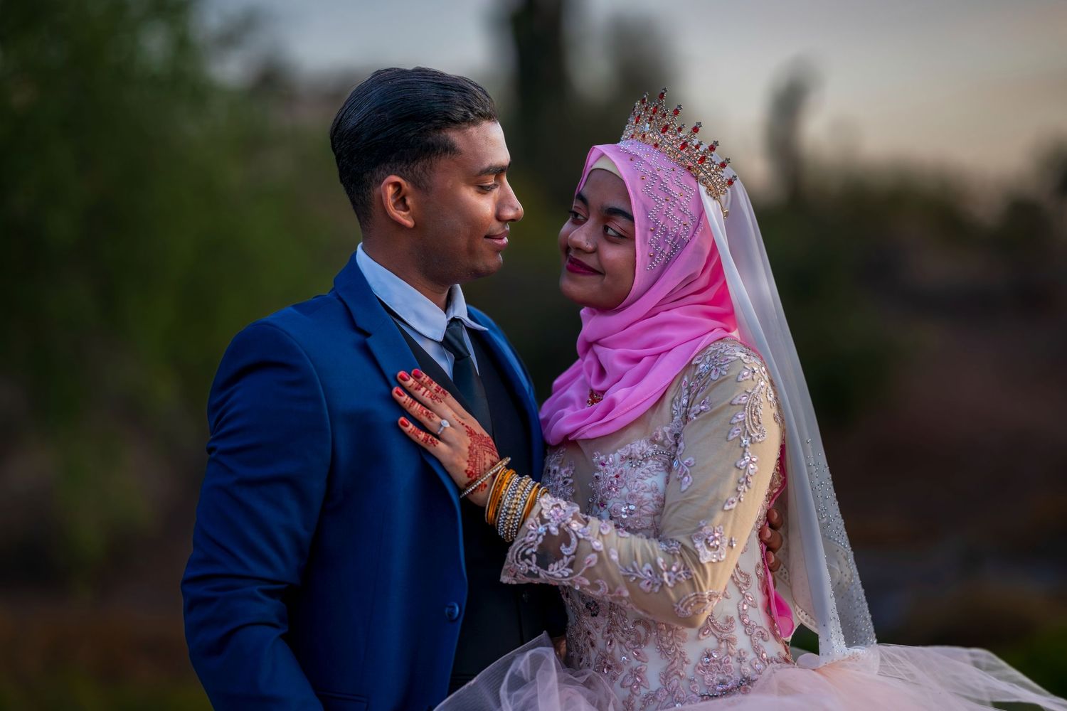 Muslim wedding couple in blue suit and pink hijab bridal gown sharing romantic moment outdoors.