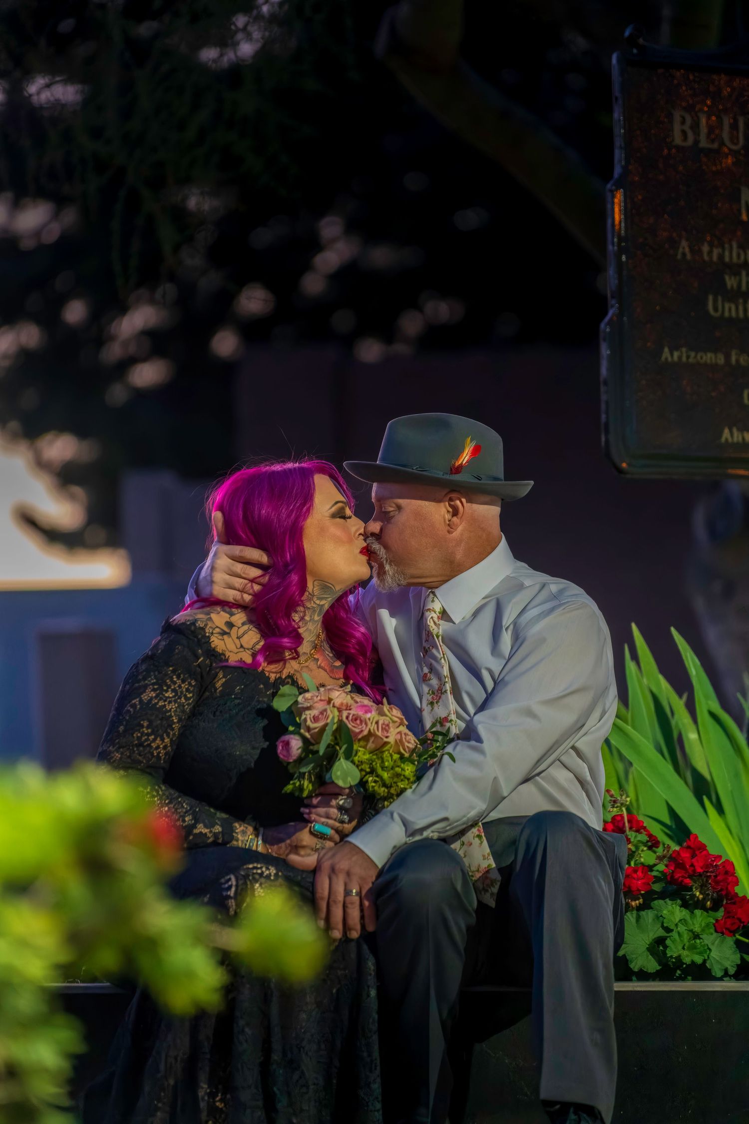 Couple sharing a romantic kiss outdoors at night, woman with pink hair holding flowers, man in fedora hat.
