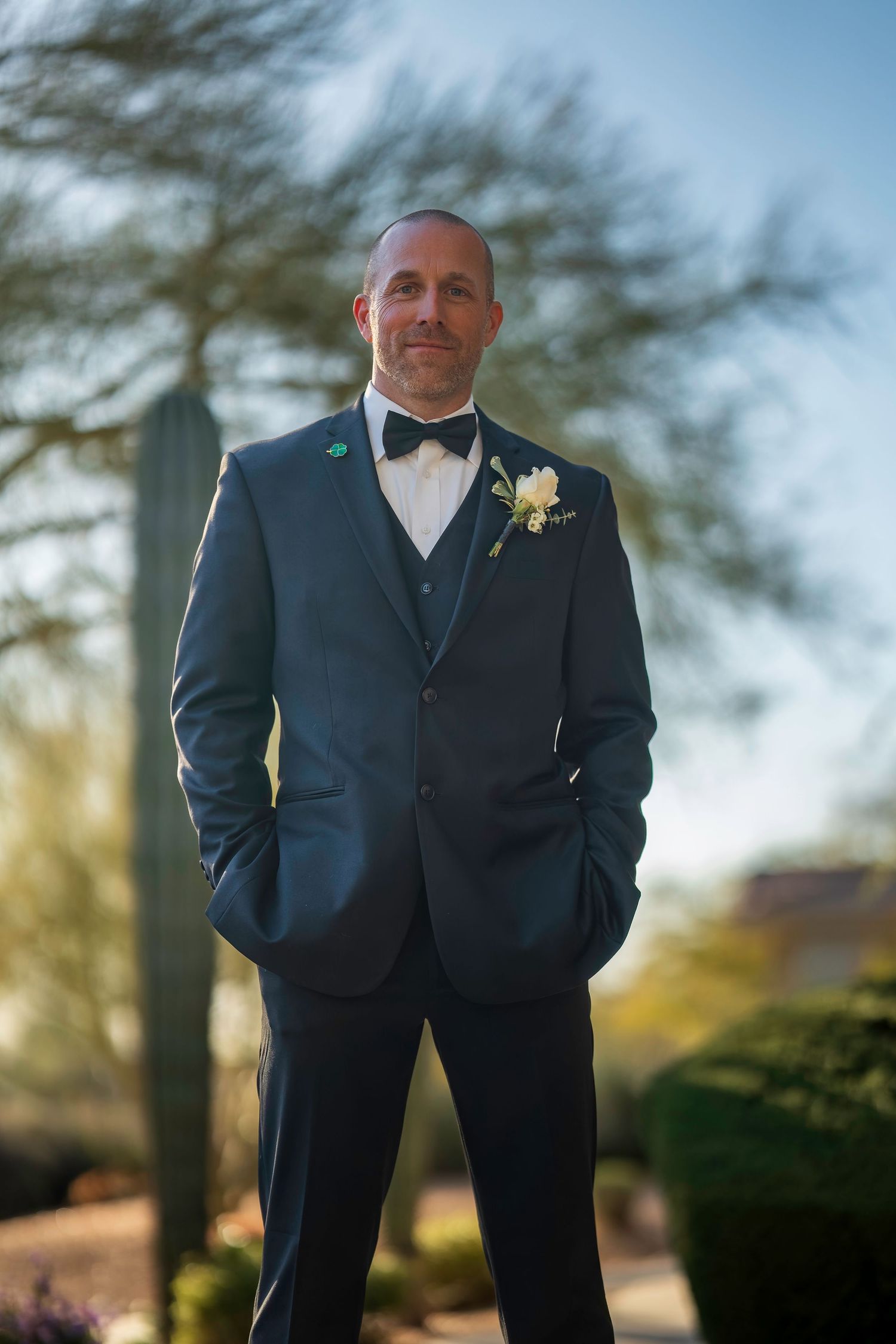 Groom in navy three-piece suit with bow tie and boutonniere poses outdoors at desert wedding venue.