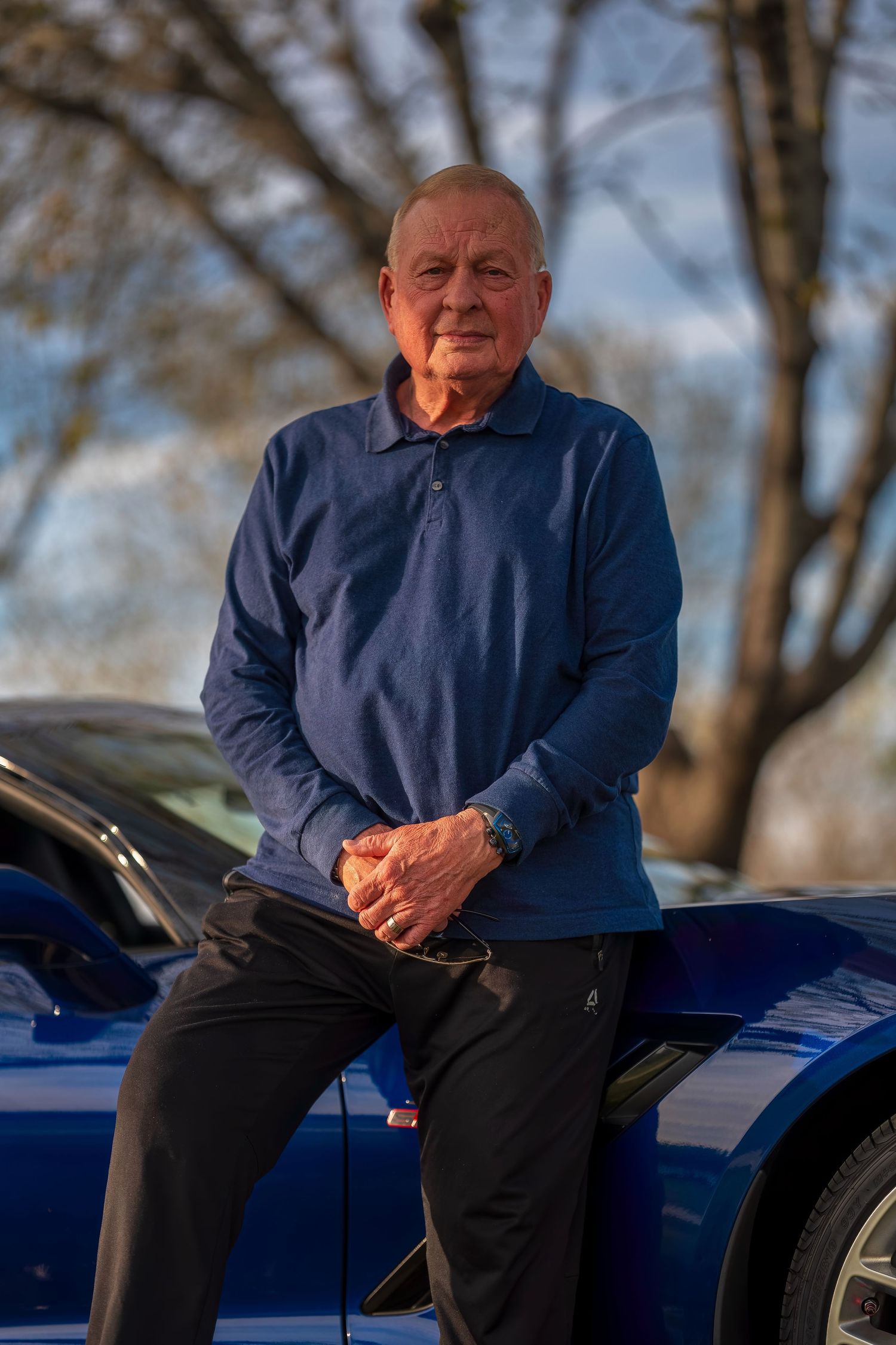 Senior man in blue polo shirt sits on hood of blue Corvette with hands clasped in outdoor setting.