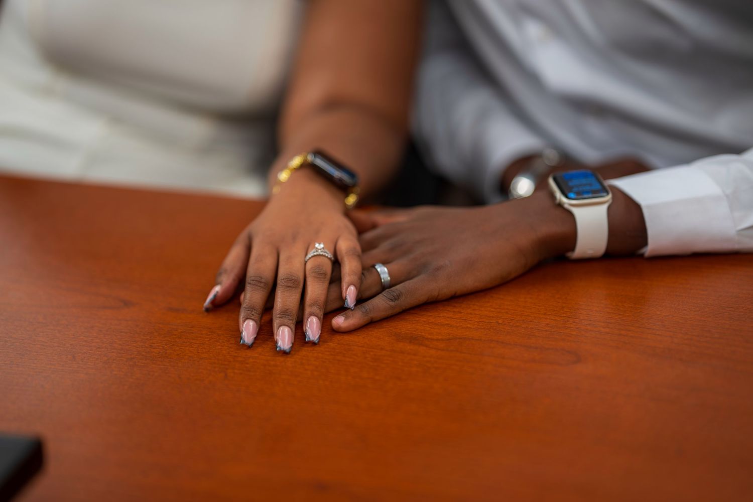 Two people holding hands on a wooden table, wearing rings and smartwatches, symbolizing love and commitment.