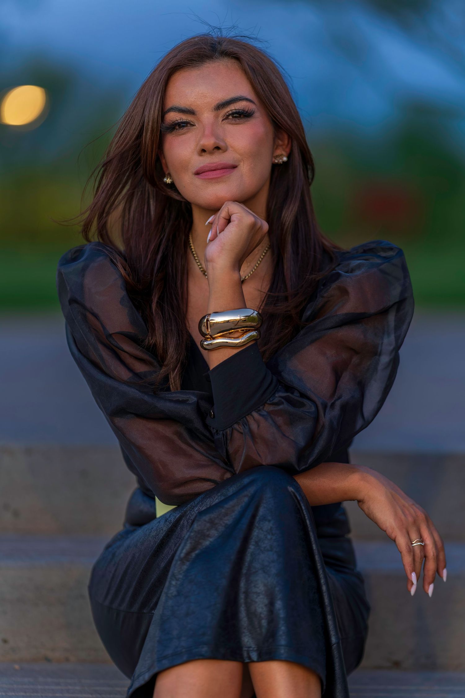 Brunette woman in black sheer puff-sleeve top and leather skirt sitting on steps at dusk.