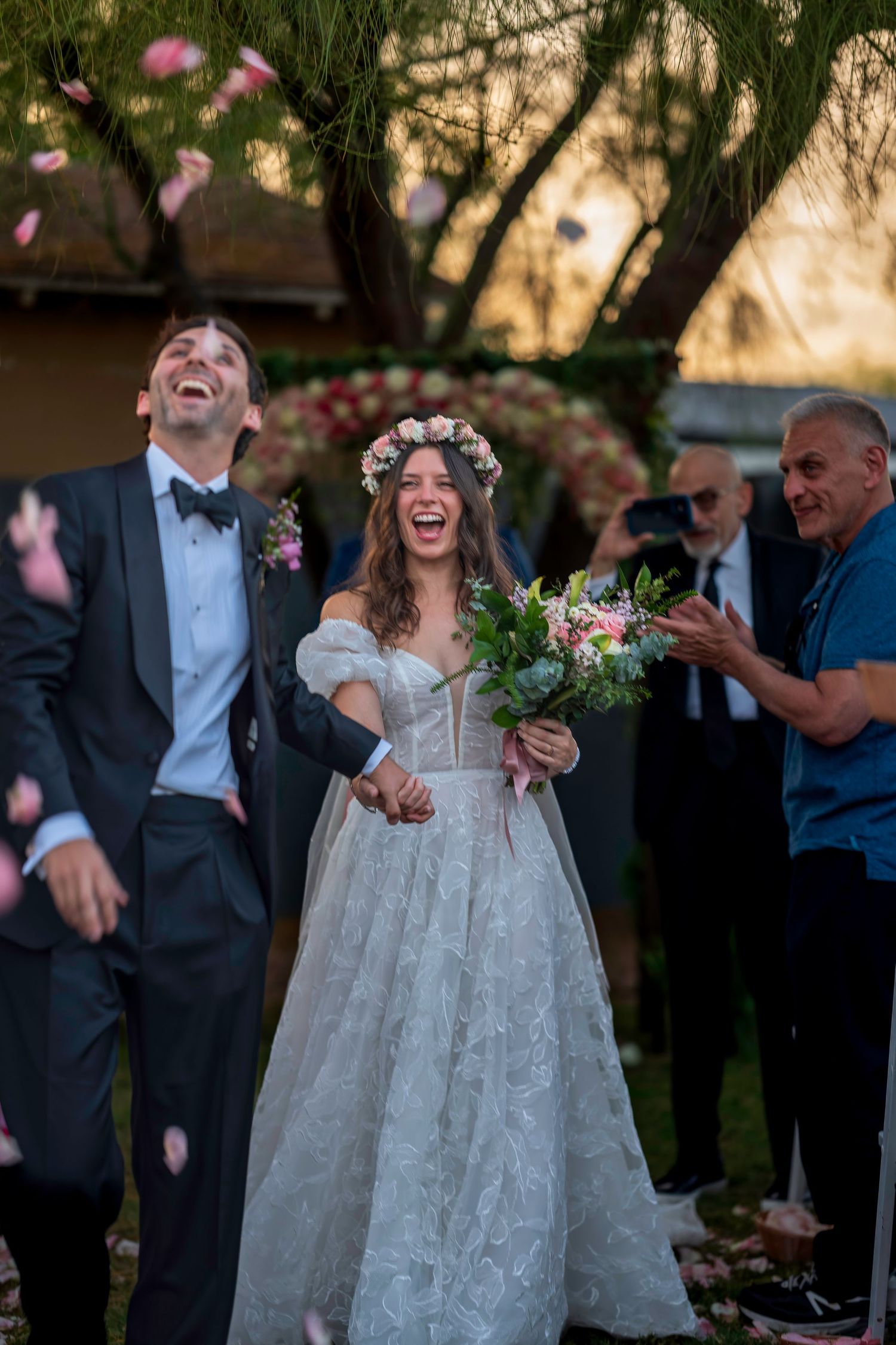 Joyful newlyweds laugh as confetti falls during outdoor wedding ceremony exit, bride in floral crown.