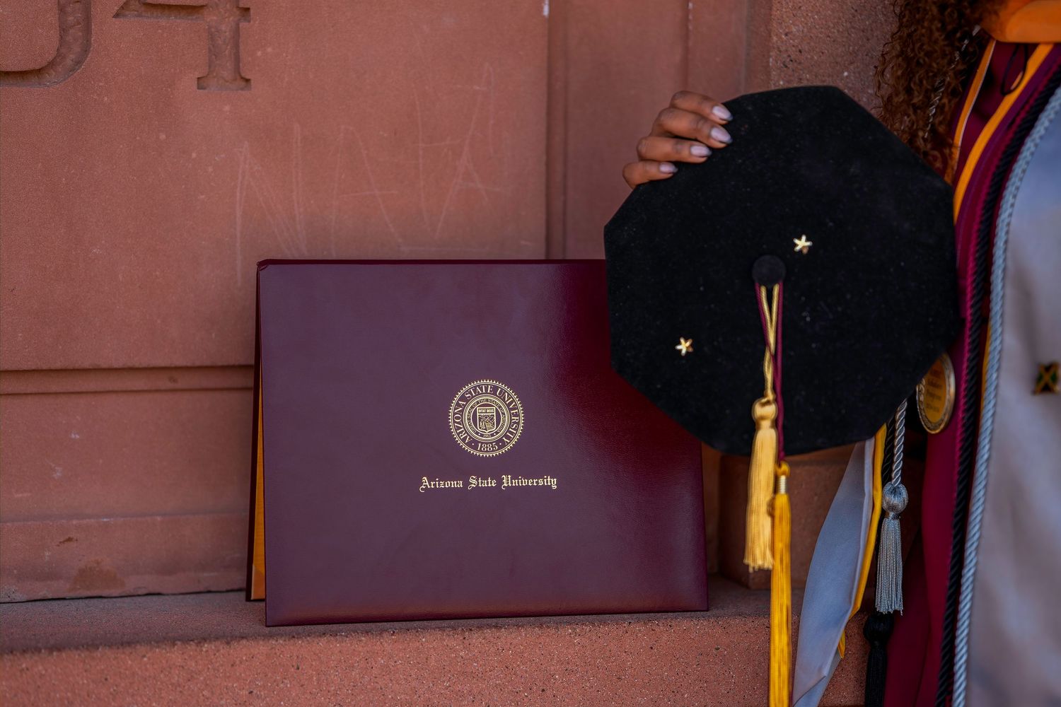 Arizona State University diploma cover held beside a black doctoral graduation cap with gold stars and yellow tassel.