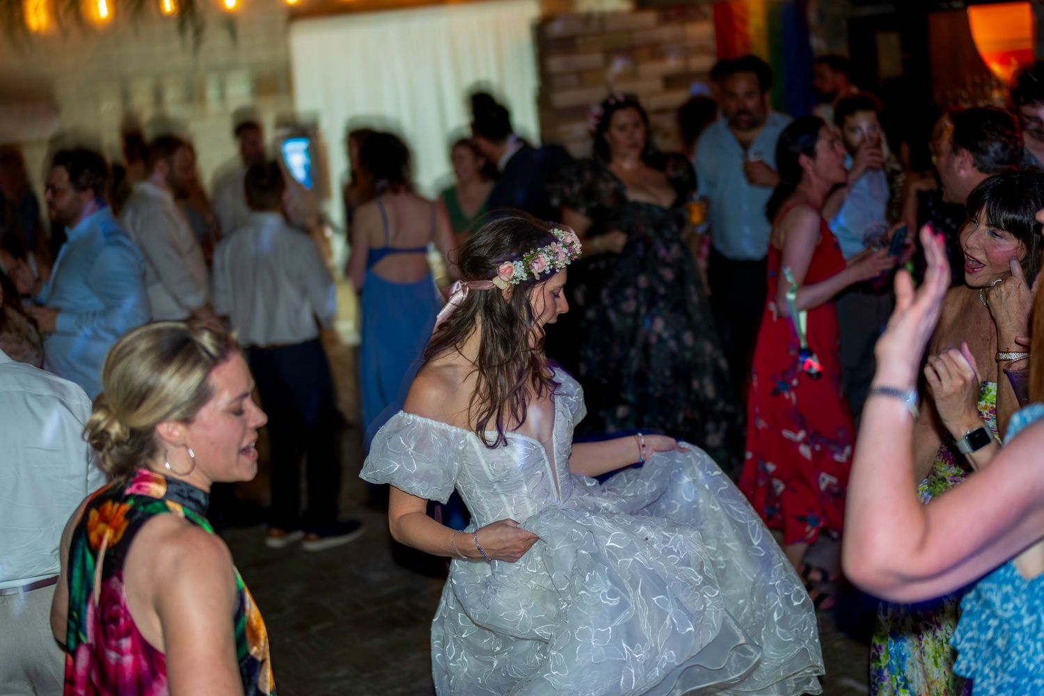 Bride in white lace gown and floral crown dancing joyfully with guests at a lively wedding reception.