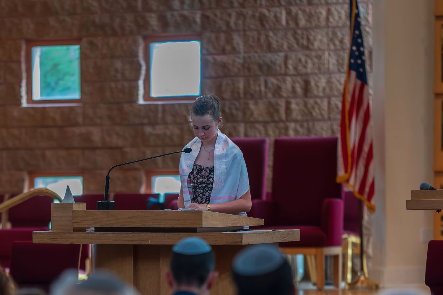 Woman speaking at podium in synagogue with American flag, wearing tallit during Jewish religious service.