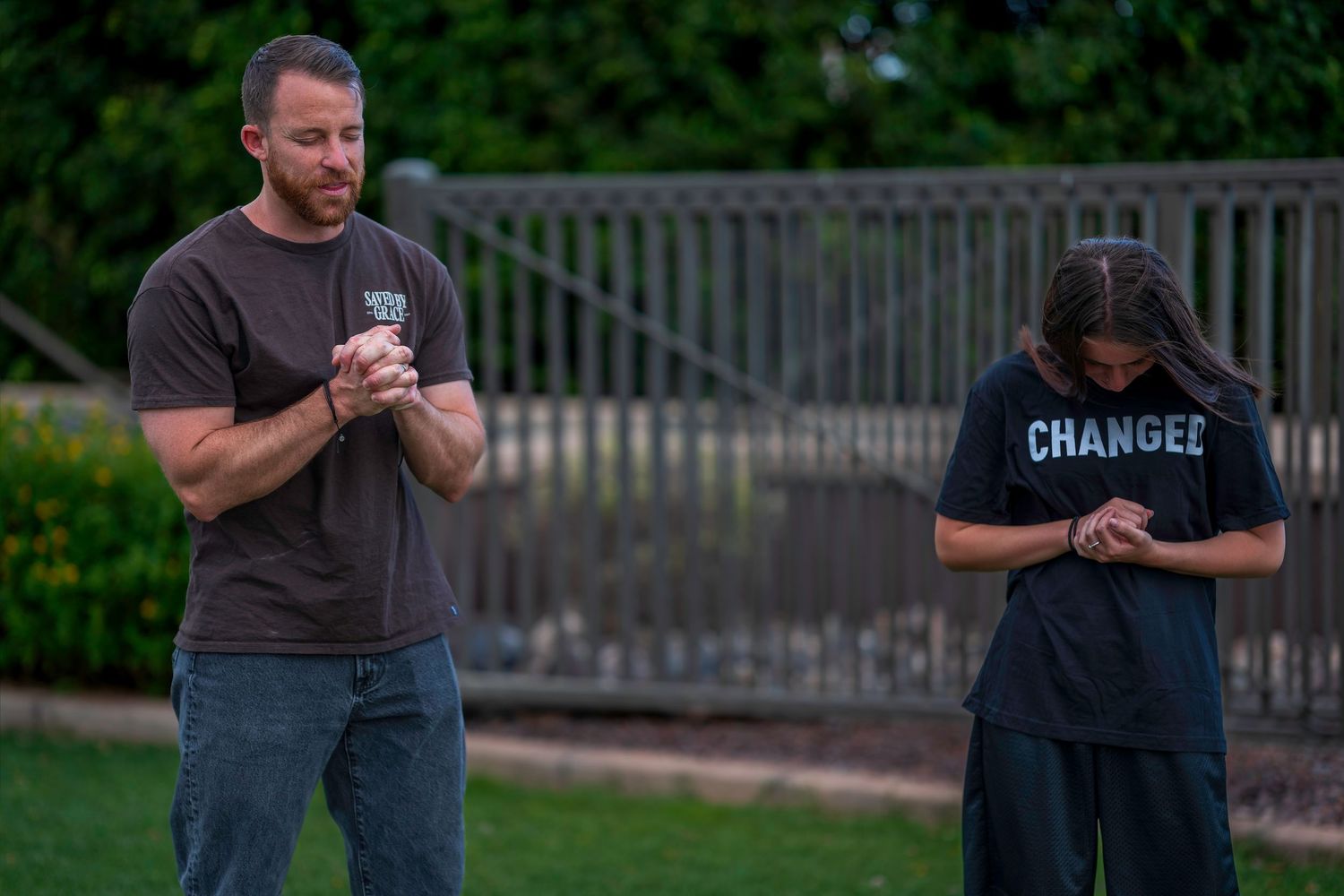 Two men standing outdoors bowing heads in prayer, one wearing a black Change shirt.