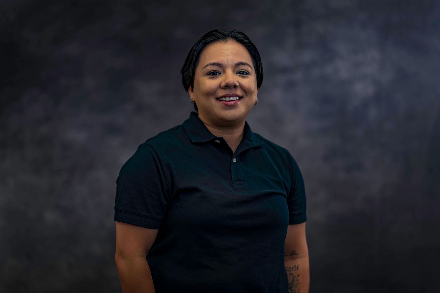 Staff member in dark polo shirt poses for company headshot against gray background.