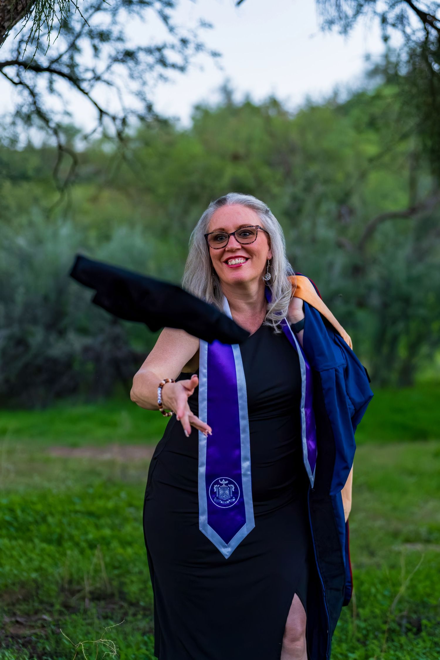 Graduate in blue academic regalia with purple stole poses joyfully in outdoor setting.