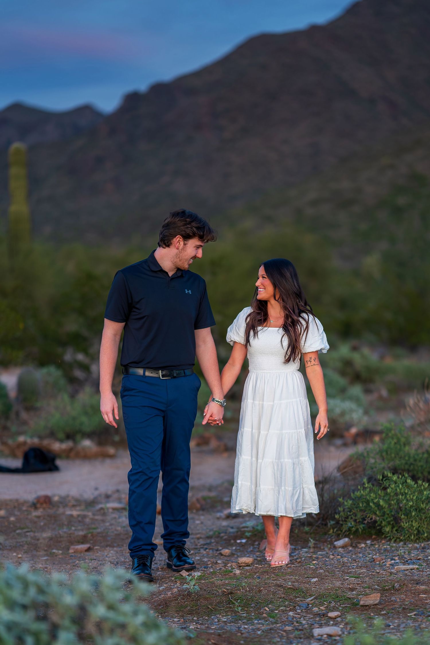 A couple walking together in a desert setting with mountains and cacti in the background.