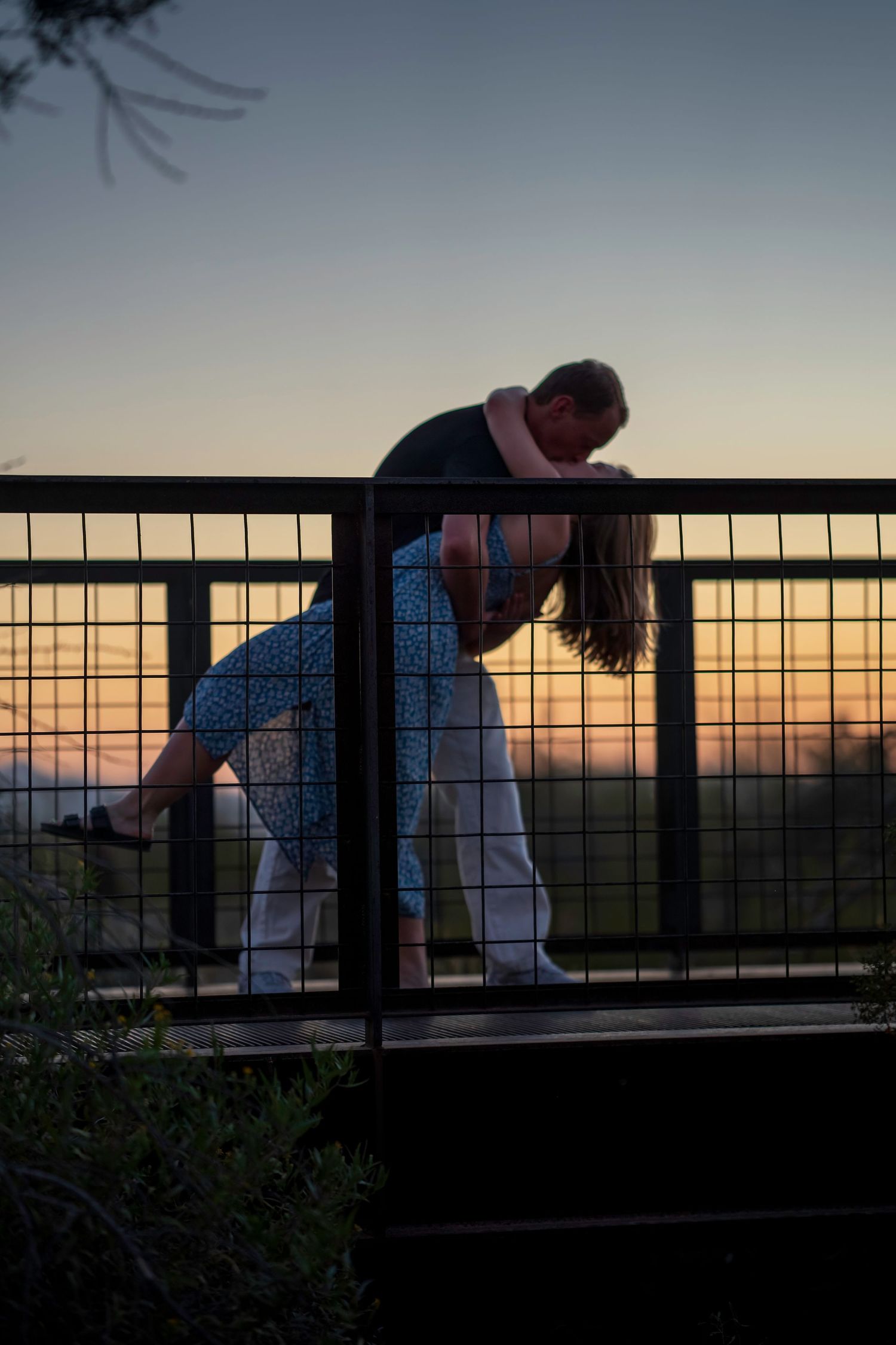 Romantic silhouette of couple sharing a dip kiss on bridge railing against colorful desert sunset sky.