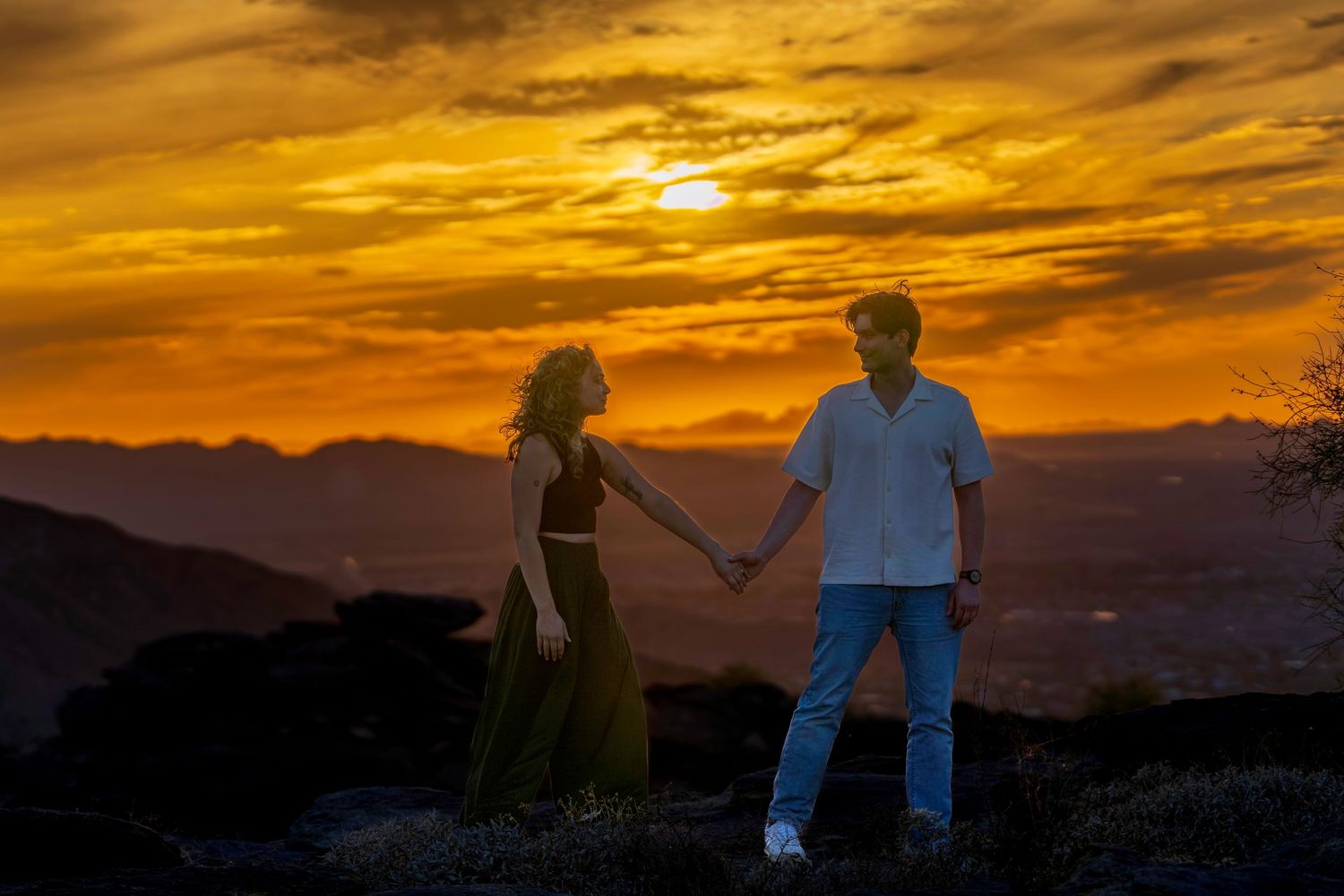 Couple holding hands at golden sunset with mountain silhouettes and vibrant orange sky backdrop.