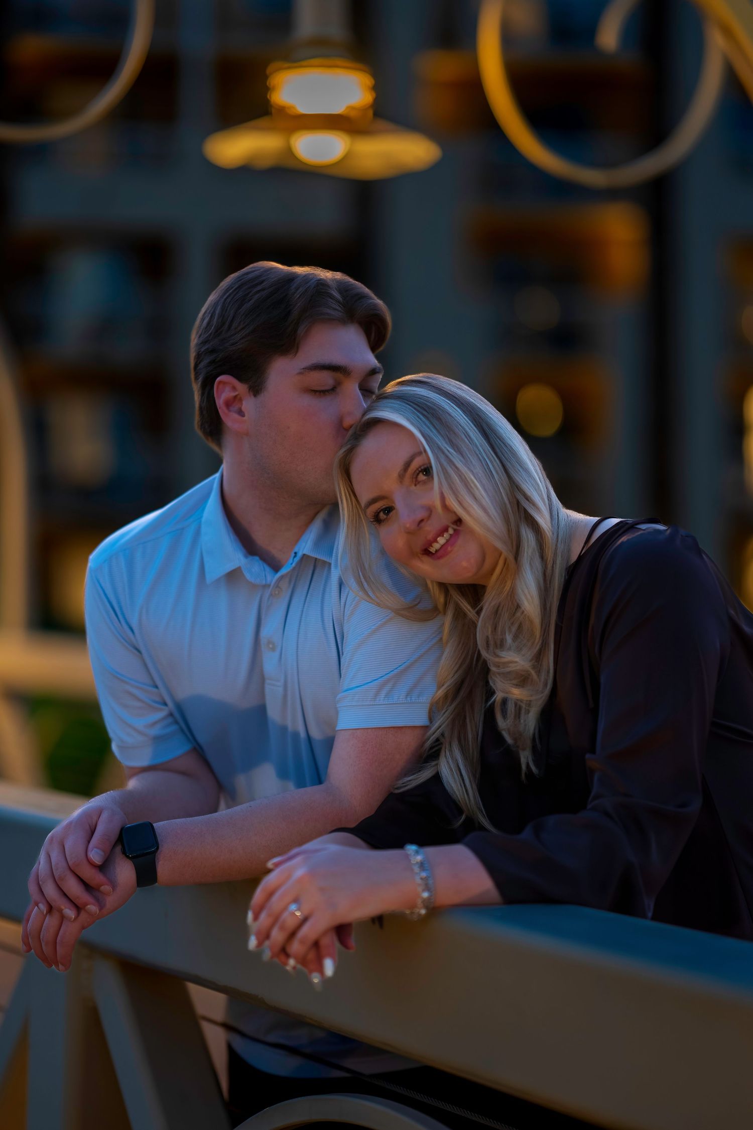 Man kissing woman on cheek on illuminated bridge at night, couple leaning on railing smiling.