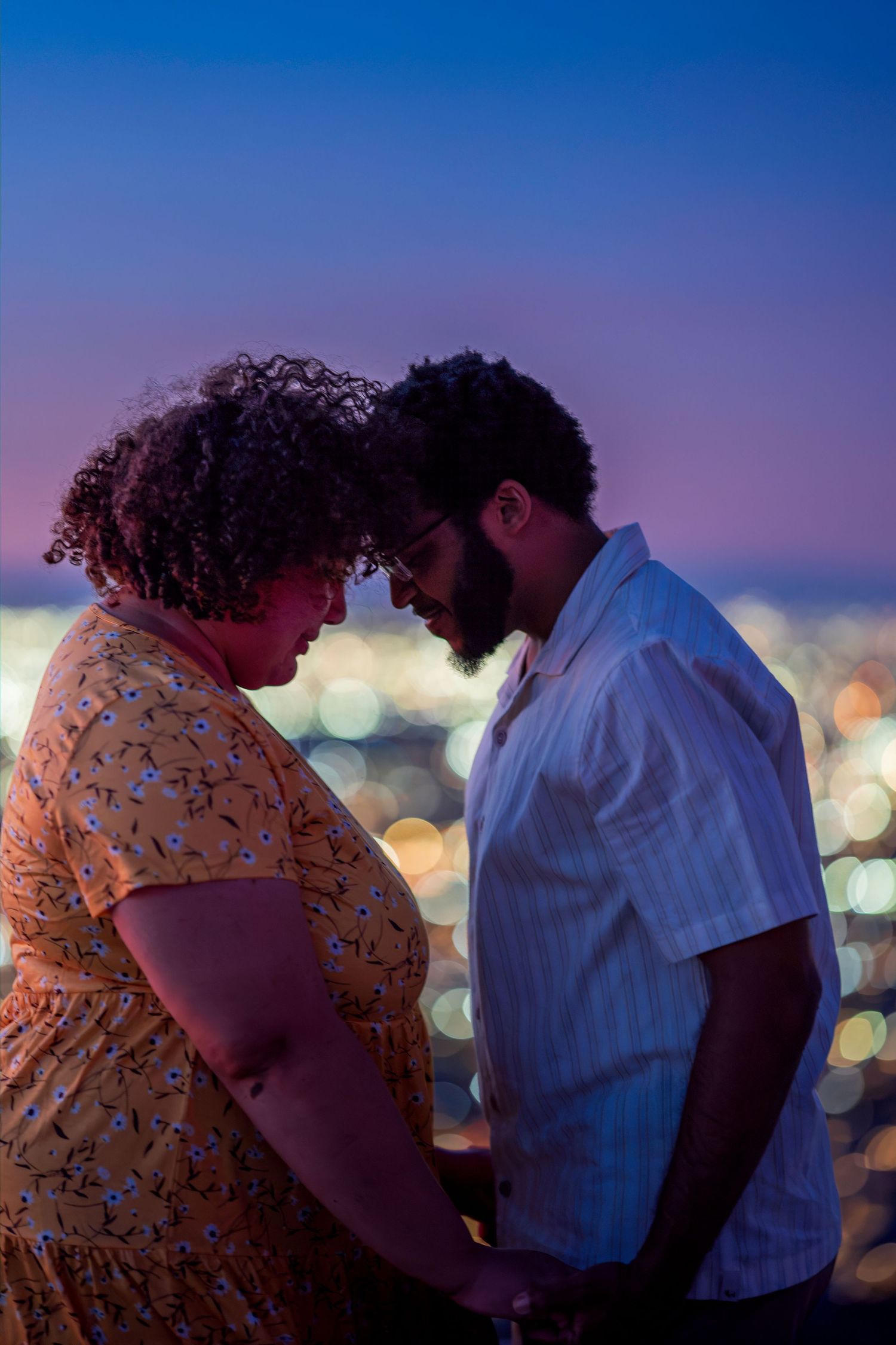 Couple holding hands and touching foreheads tenderly with bokeh city lights at night.