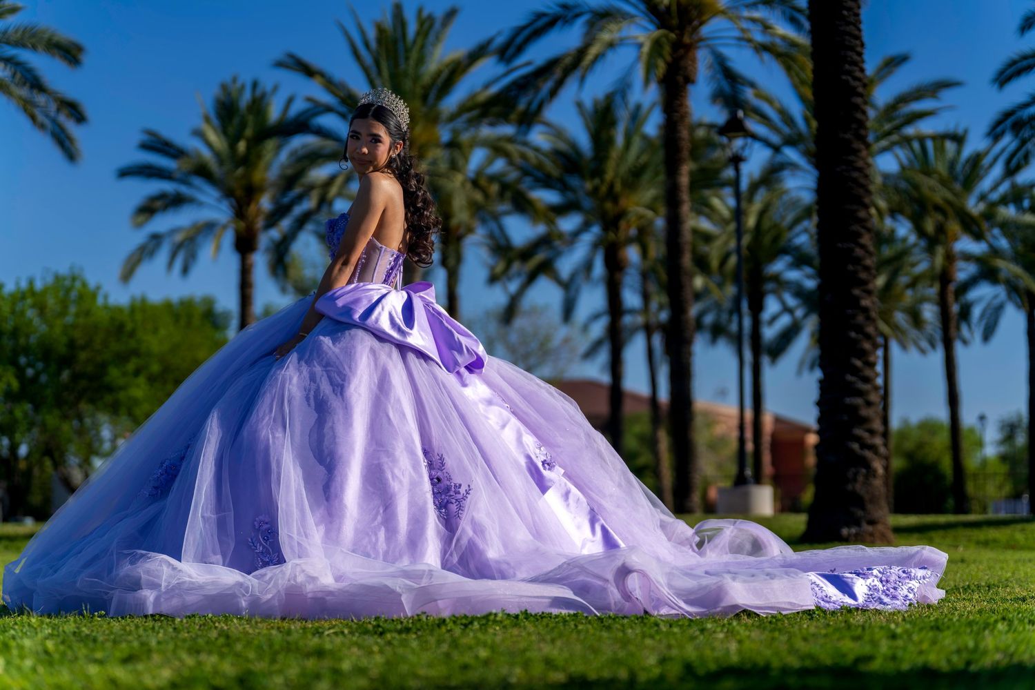 Young woman in lavender quinceañera ball gown poses outdoors with palm trees and blue sky backdrop.