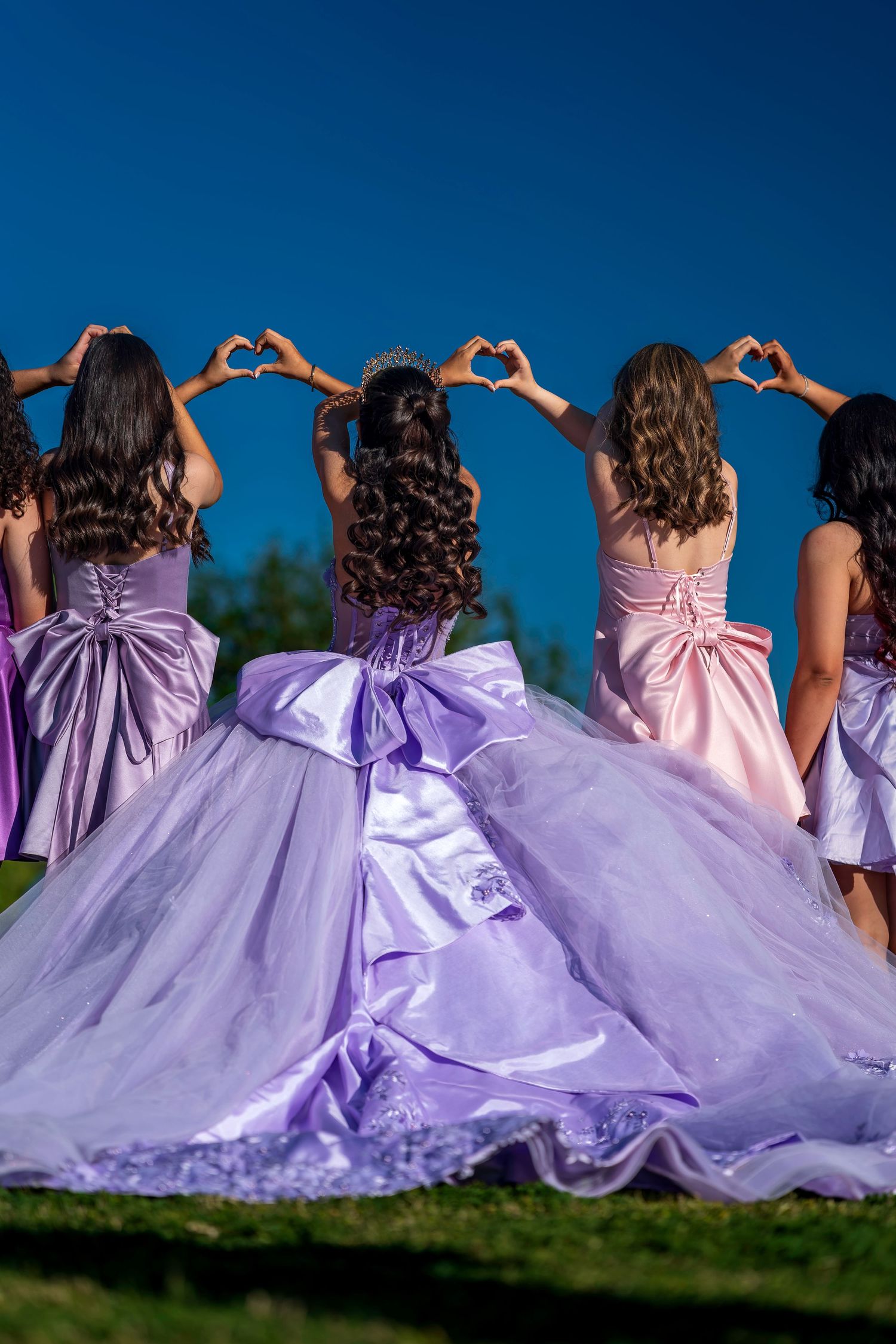 Five girls in purple and pink ball gowns stand with backs turned, making heart shapes with their hands against a blue sky.
