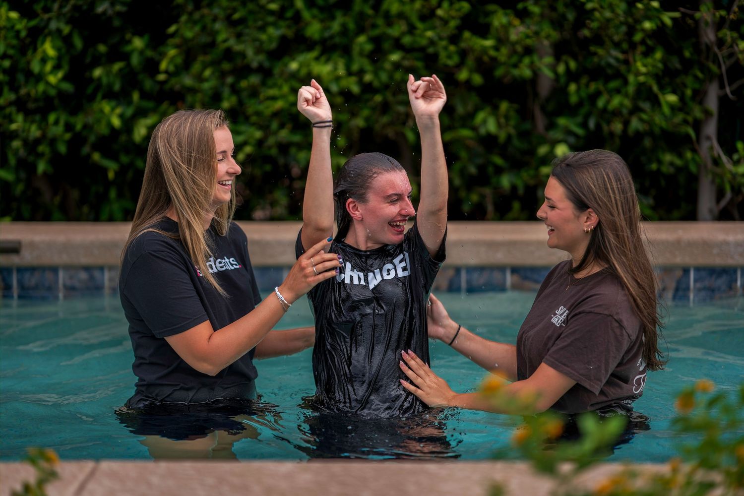 Three women in wet black shirts celebrate a baptism in an outdoor pool, with arms raised in joy.