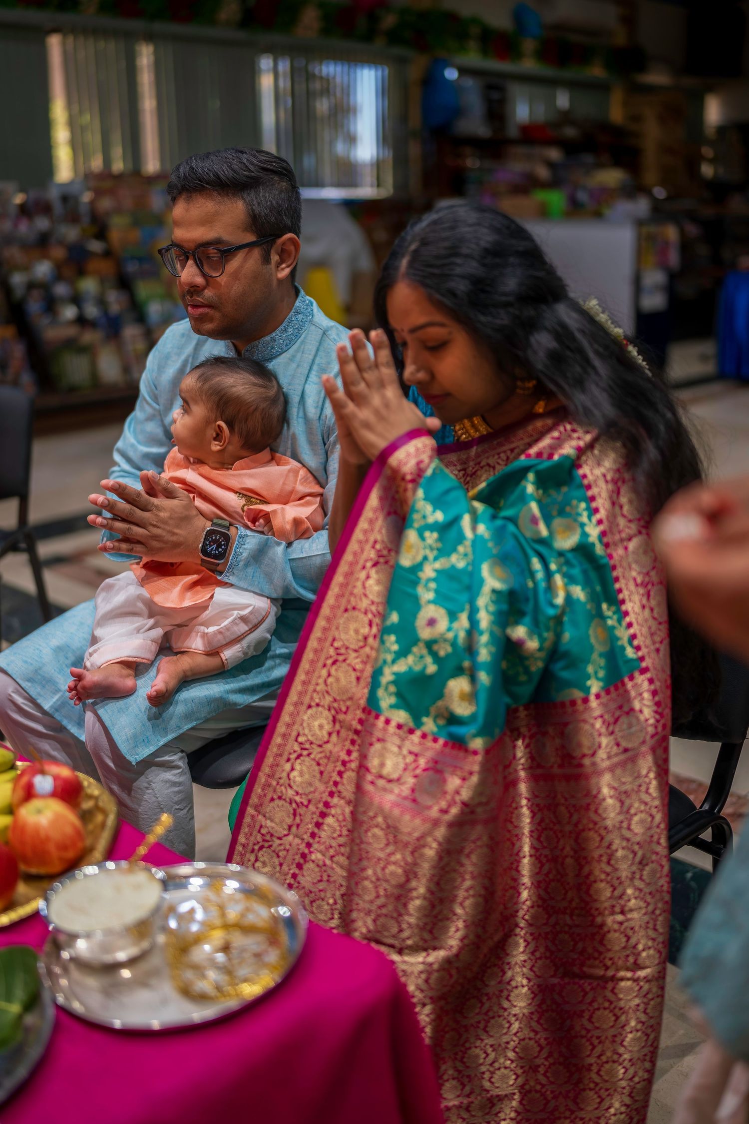 Indian family at religious ceremony, man holding baby while woman prays in teal and gold saree near offerings.