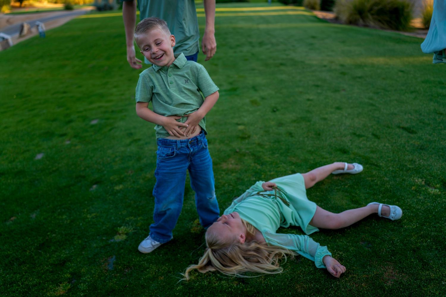 A laughing boy stands on green grass while a girl with long blonde hair lies dramatically on the ground.