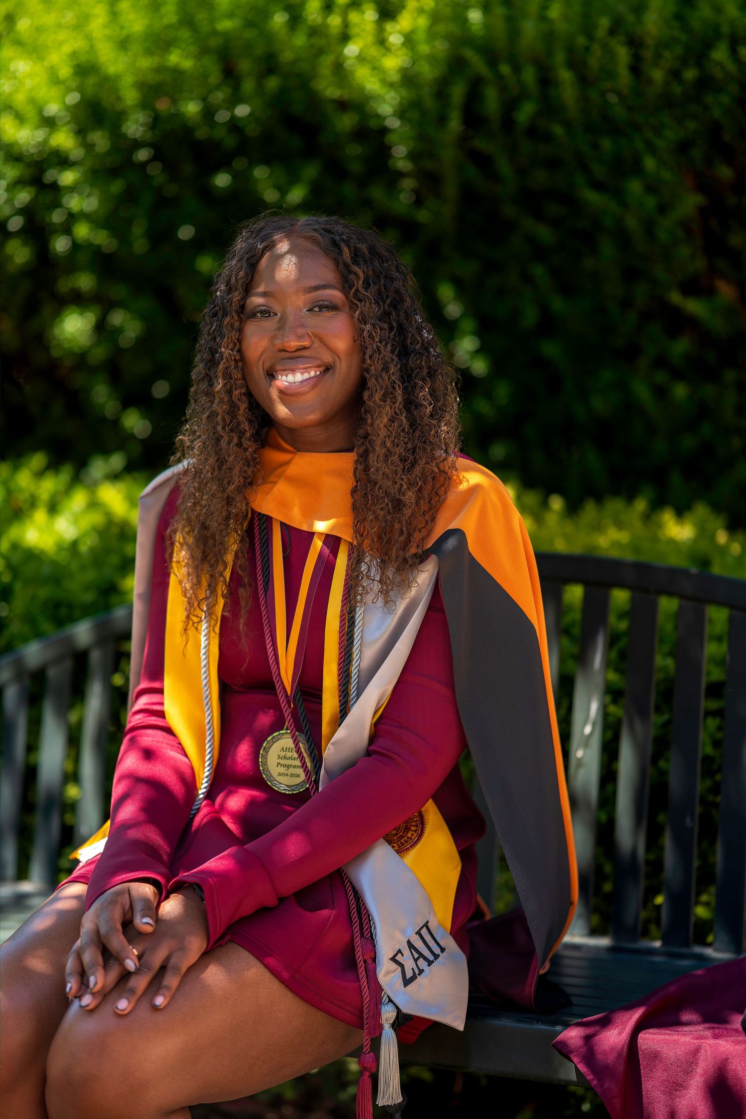 Smiling female graduate in maroon and gold ASU regalia with Sigma Alpha Pi stole seated outdoors.