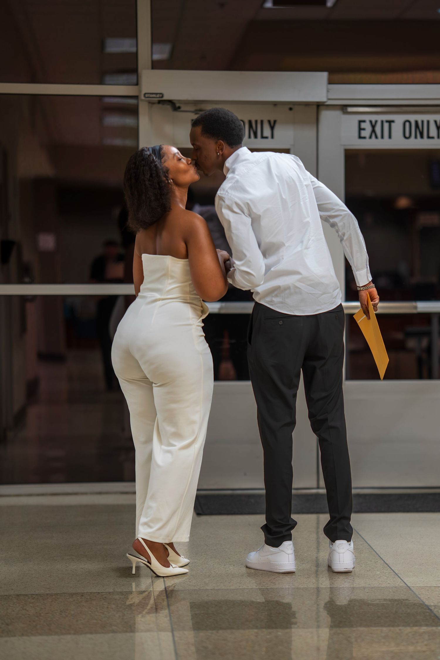 A newlywed couple shares a kiss near exit doors, bride in white jumpsuit and groom holding documents.