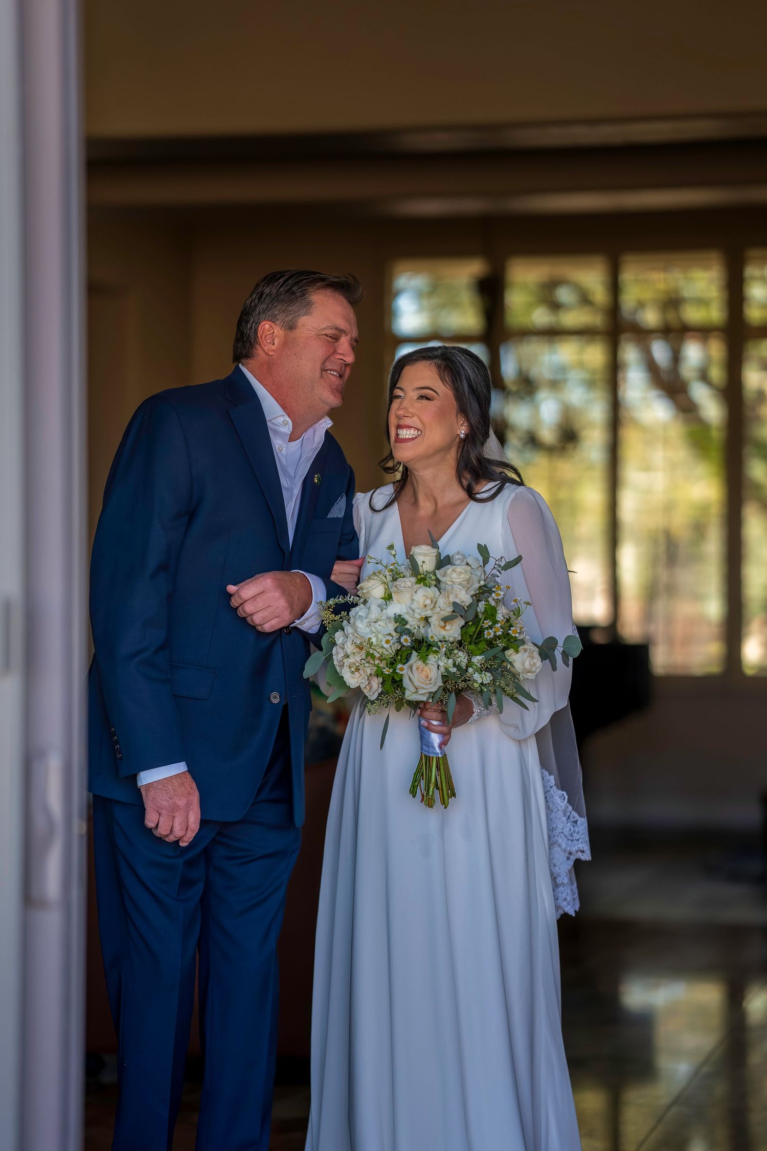Bride in white gown holding bouquet smiles at man in blue suit during indoor wedding ceremony.