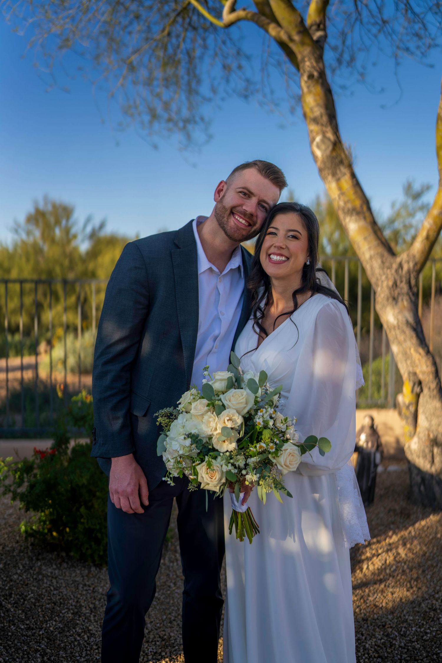 Happy bride and groom embracing outdoors holding white rose bouquet at sunset wedding ceremony.