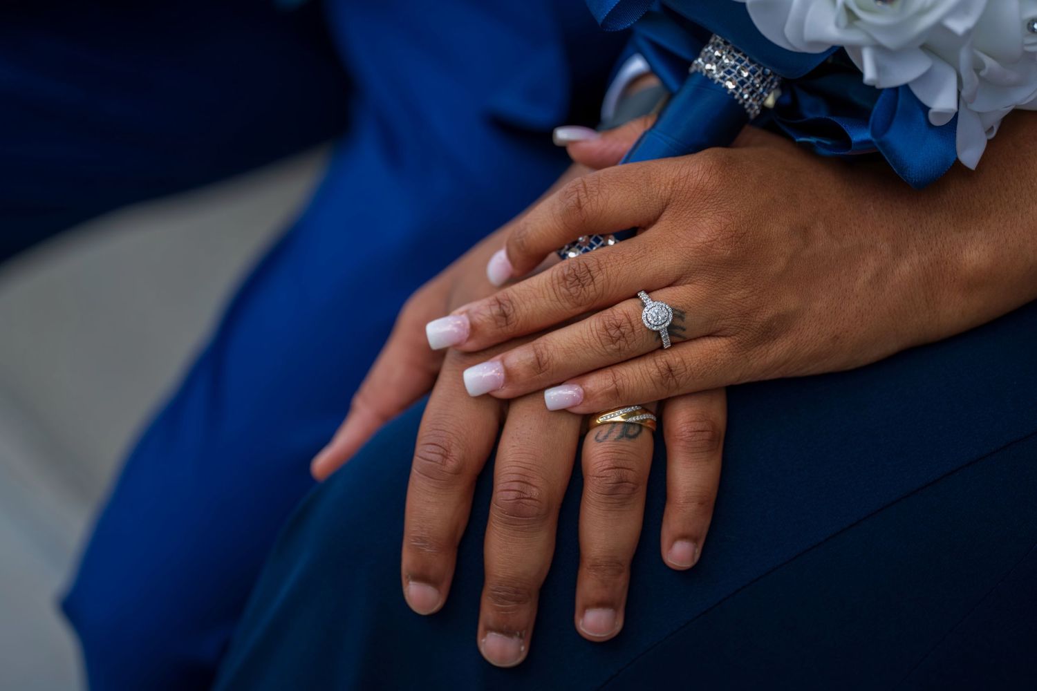 Newlywed couple displaying wedding rings and engagement ring on hands, wearing blue suit with floral bouquet.