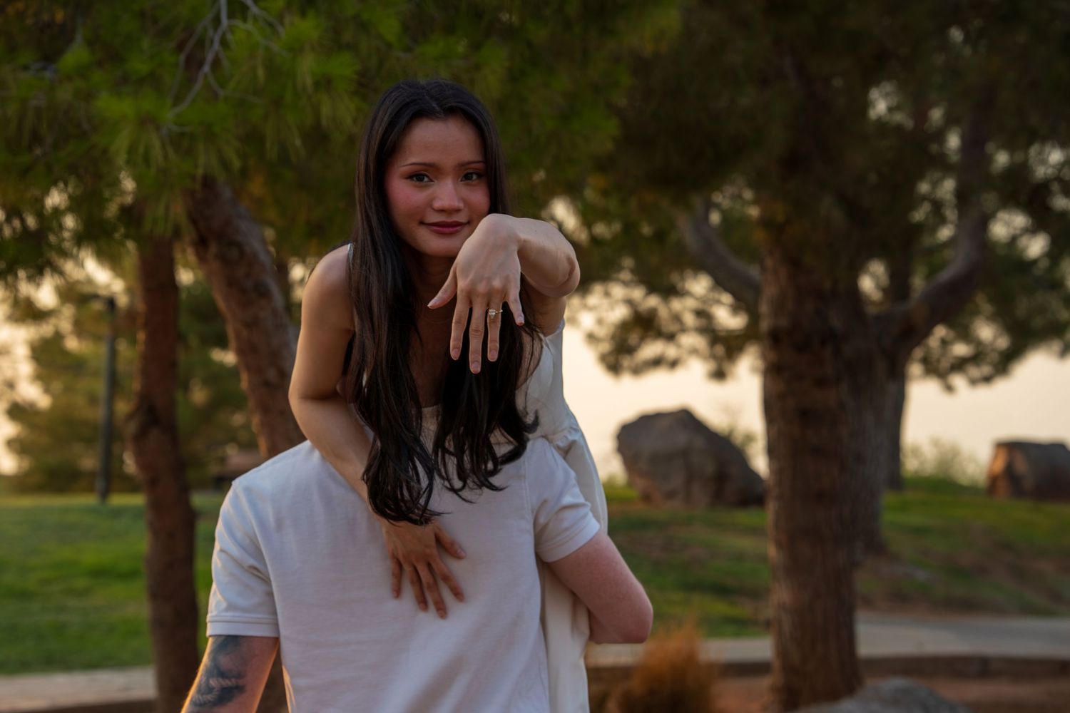 Young woman with long dark hair piggyback riding a man, showing engagement ring at sunset park.