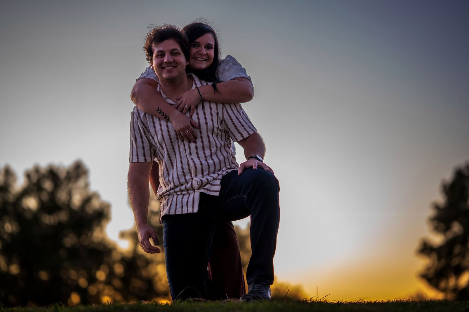 Playful couple at sunset, man kneeling on grass while woman hugs him from behind outdoors.