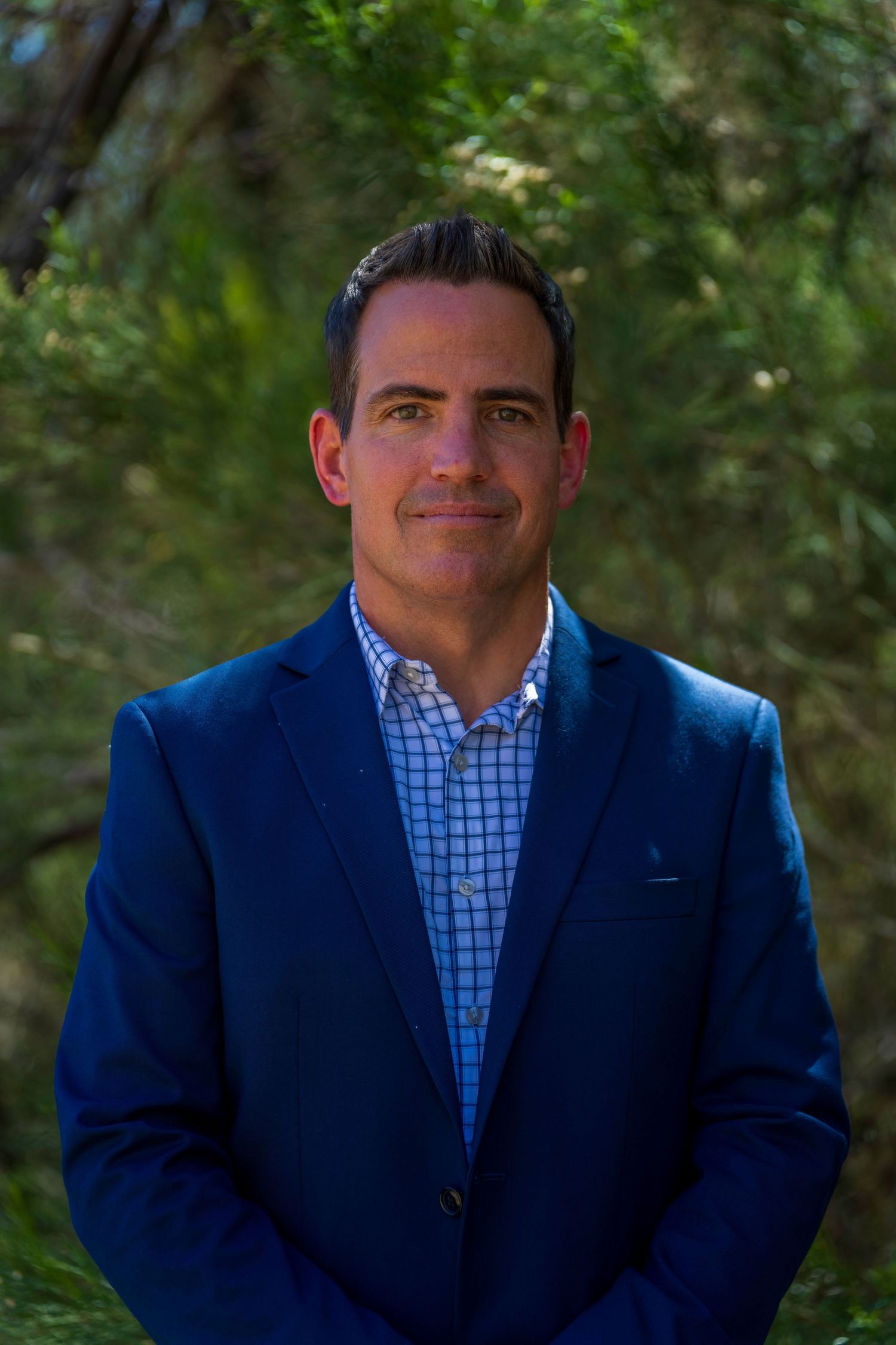 A professional man in a blue blazer and checkered shirt smiles outdoors with lush green foliage background.