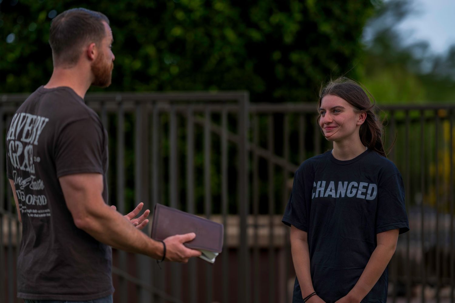 Two young men having an outdoor conversation, one wearing a 'Changed' t-shirt, standing near a metal fence.