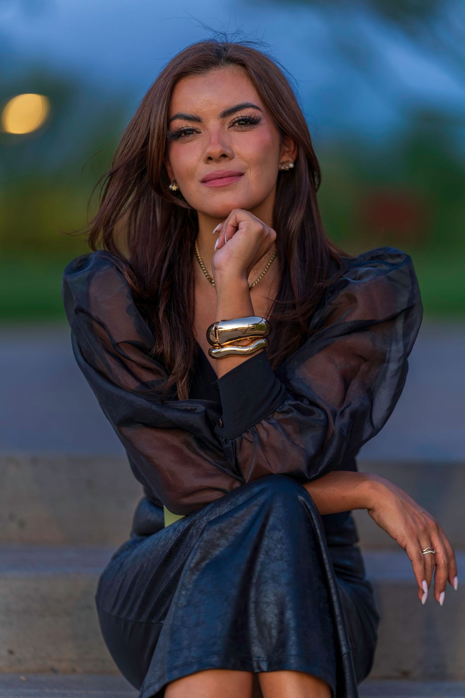 Elegant woman in black sheer blouse and leather skirt sitting on steps at dusk, wearing gold jewelry.