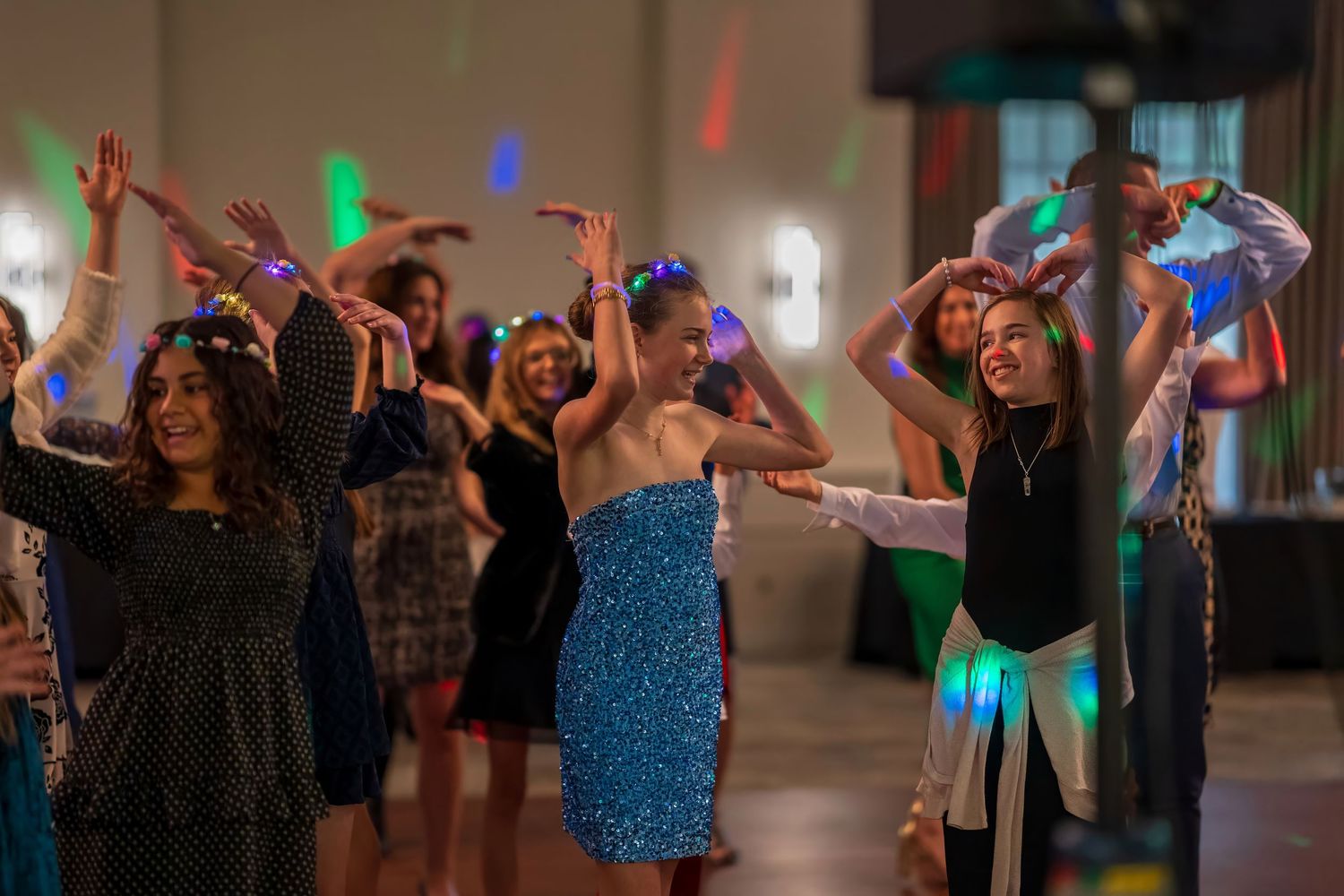 Guests dance energetically under colorful lights at a festive party or school dance event.