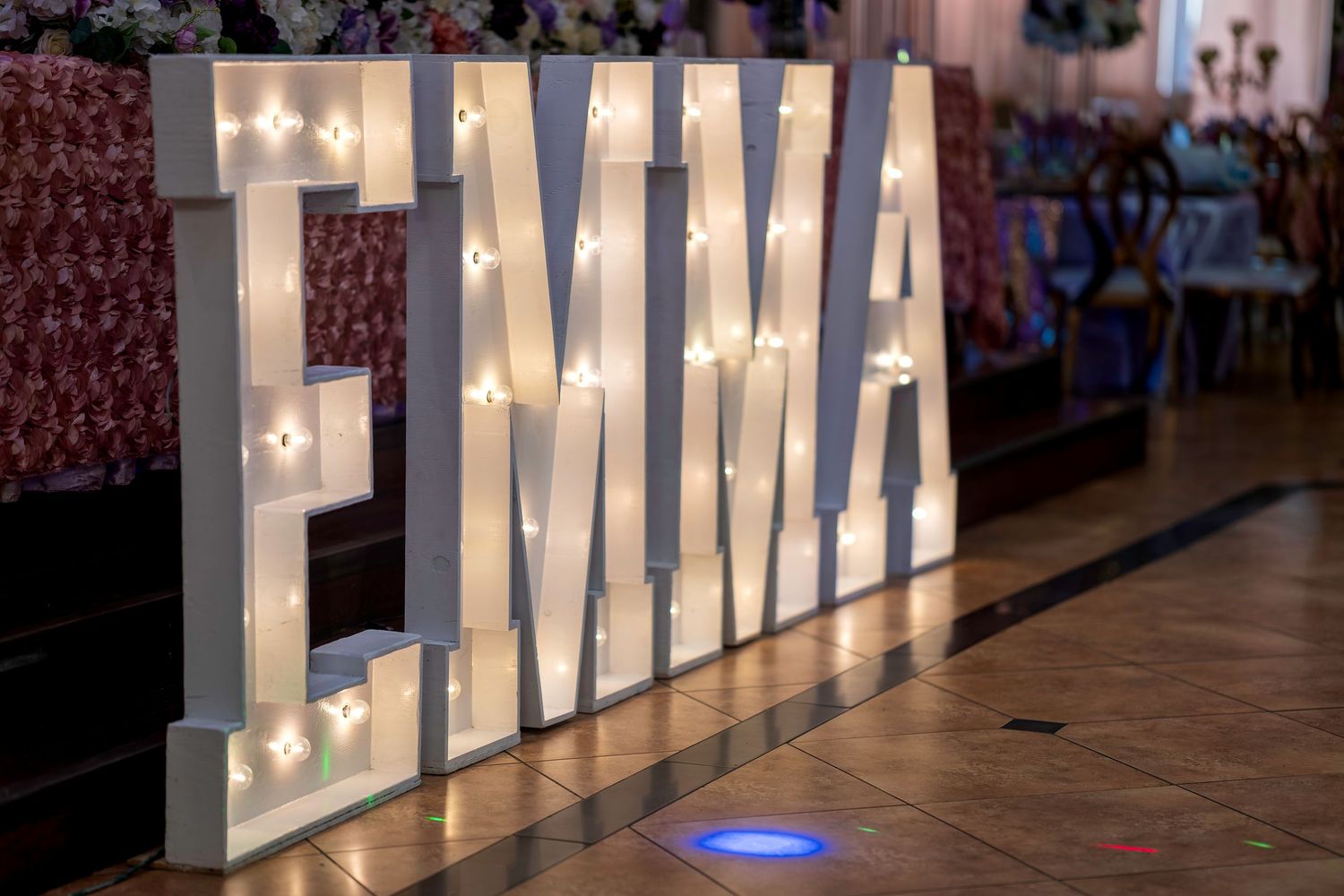 Large illuminated marquee letters spelling EMMA with warm bulb lights at an elegant event venue.