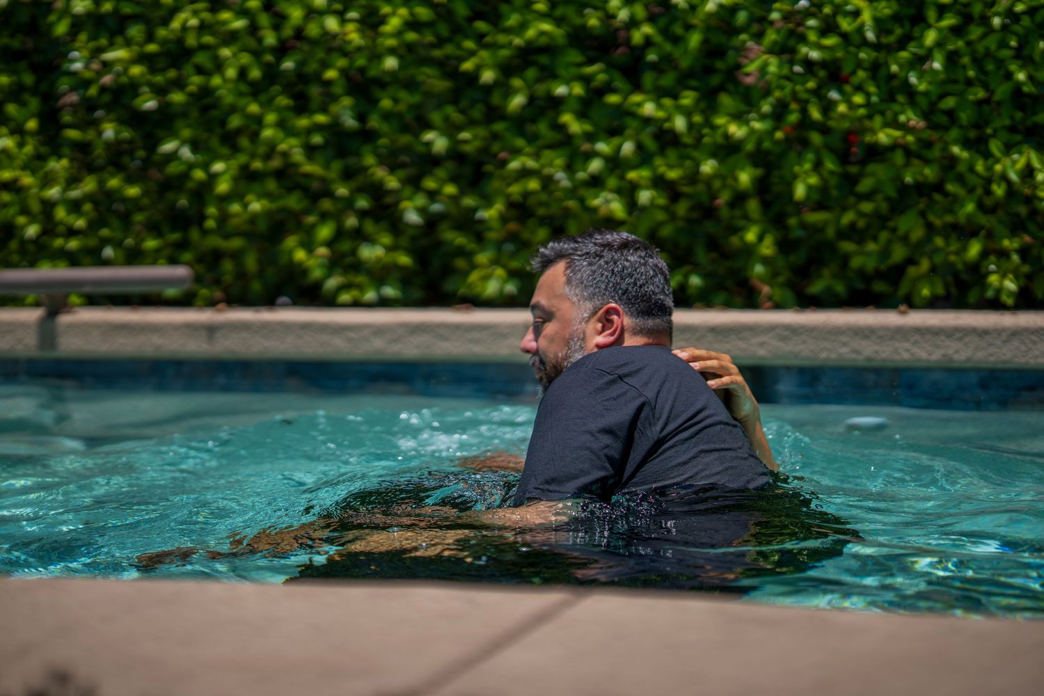 Two people embracing in a swimming pool surrounded by lush green hedges on a sunny day.