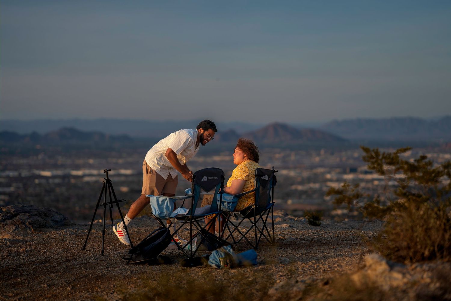 Couple enjoying a scenic overlook at dusk with camping chairs and a tripod in a desert landscape.