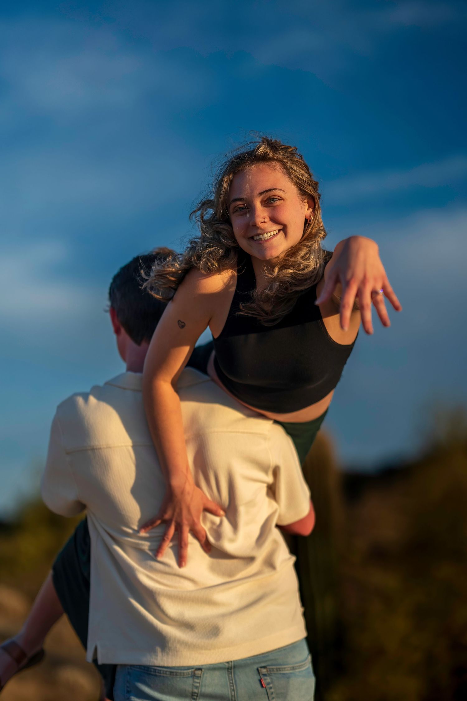 A joyful woman spreads her arms while being lifted by her partner outdoors under a blue sky.