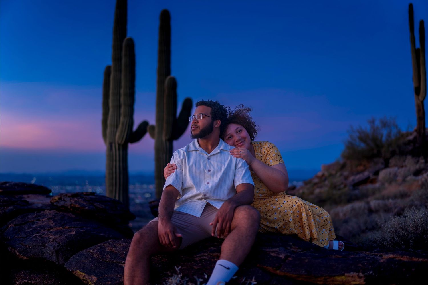 Couple sitting on rocks at twilight surrounded by saguaro cacti under a vivid blue and pink sunset sky.