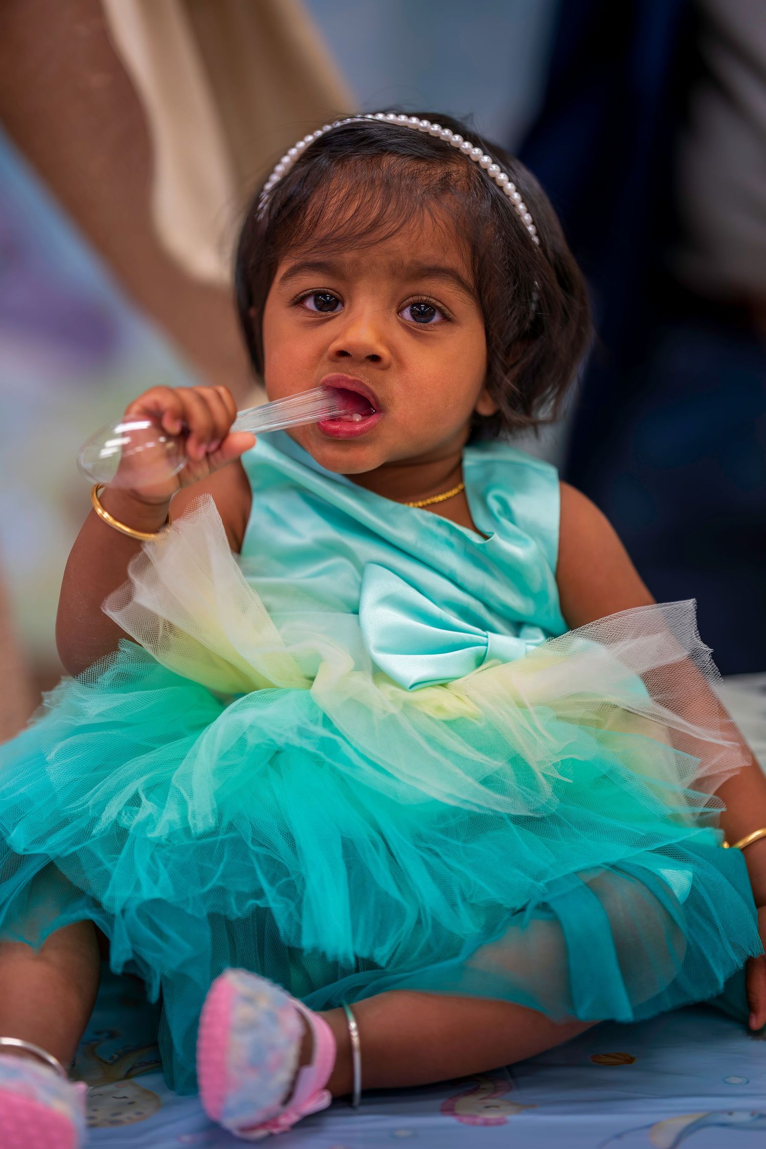 Adorable baby girl in a teal tutu dress with headband sitting on floor eating at a first birthday party celebration.