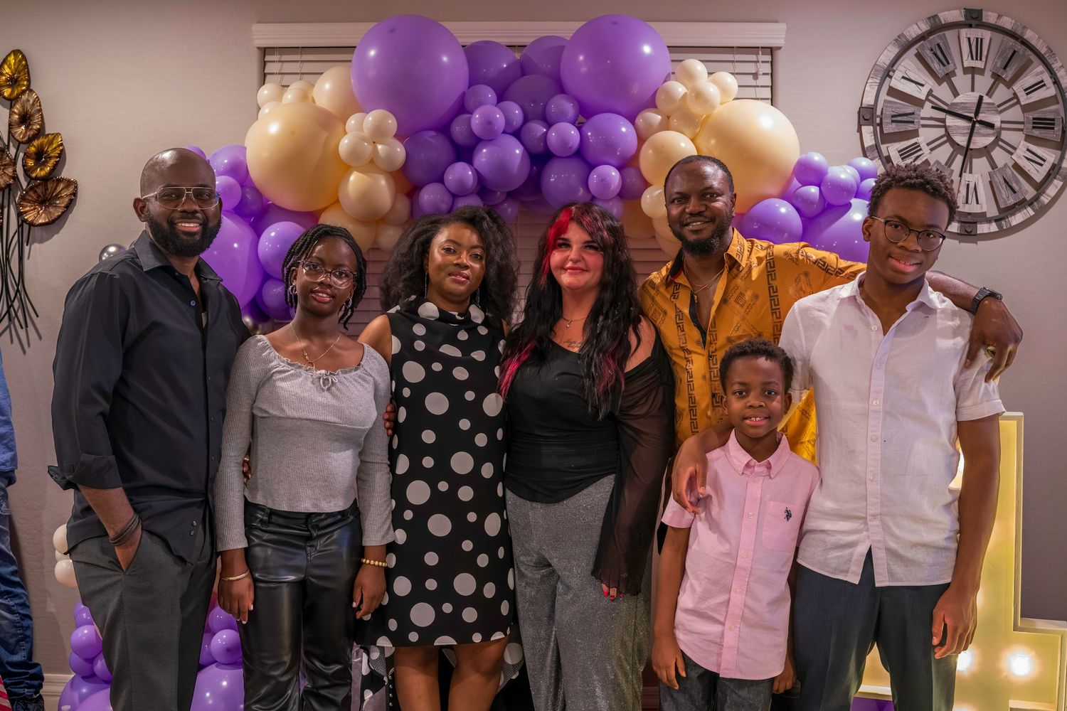Group of seven people smiling together in front of purple and gold balloon arch at a celebration event.