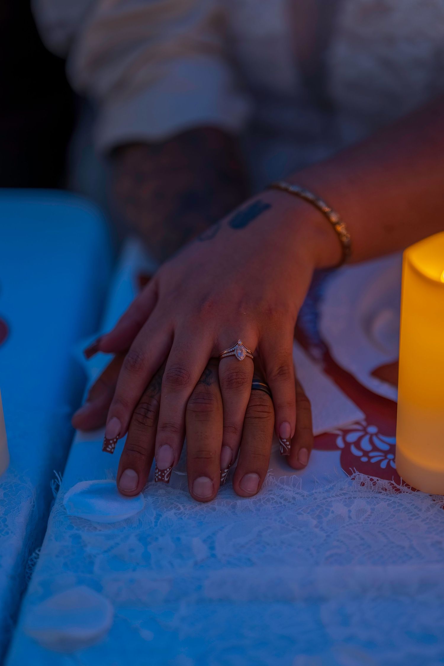Couple's hands together showing wedding rings by candlelight at reception table with lace tablecloth.