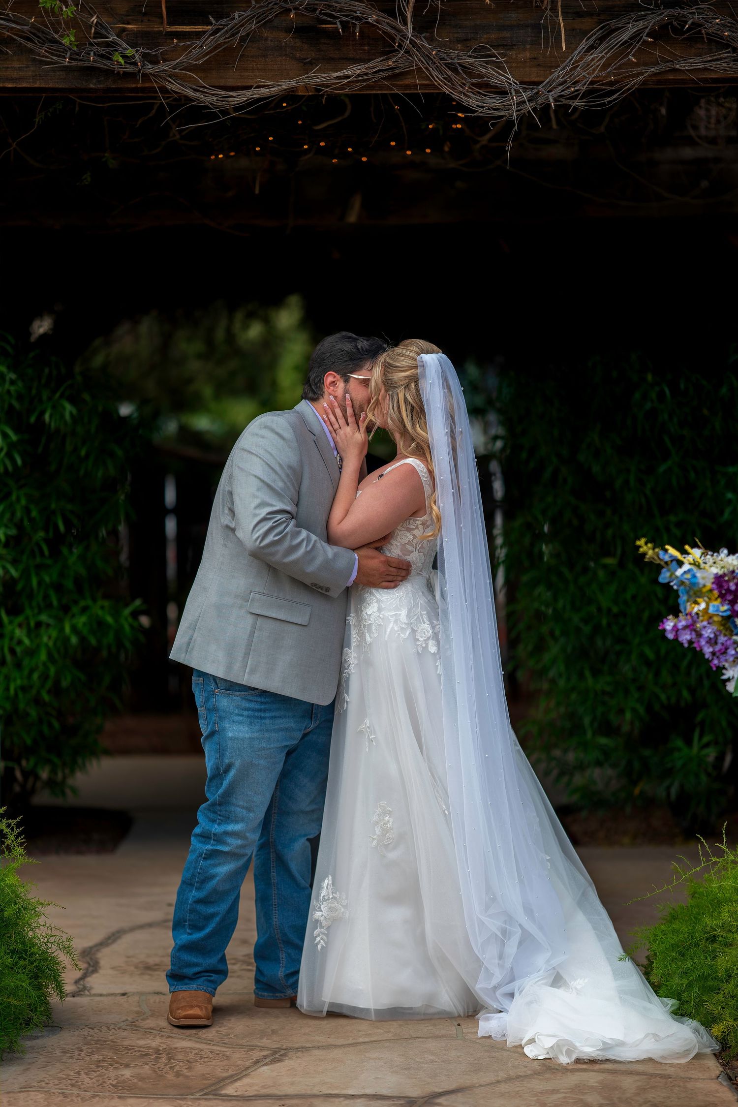 A bride and groom share their first kiss at an outdoor wedding ceremony surrounded by lush greenery.