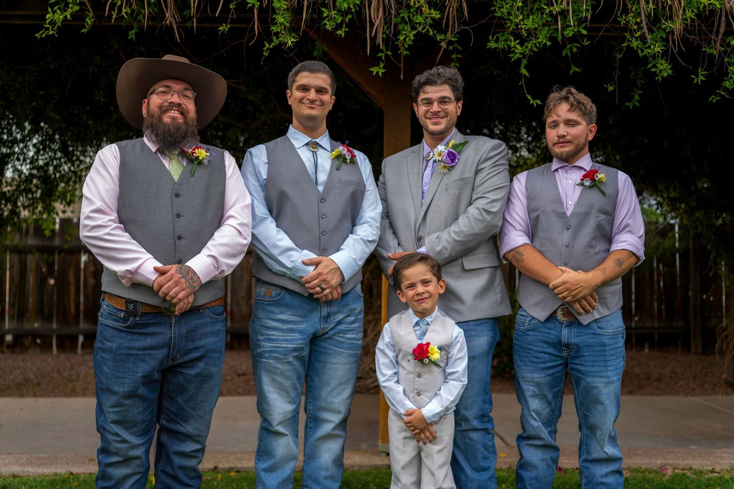 Four groomsmen and a young ring bearer posing outdoors in gray vests and jeans for a wedding photo.