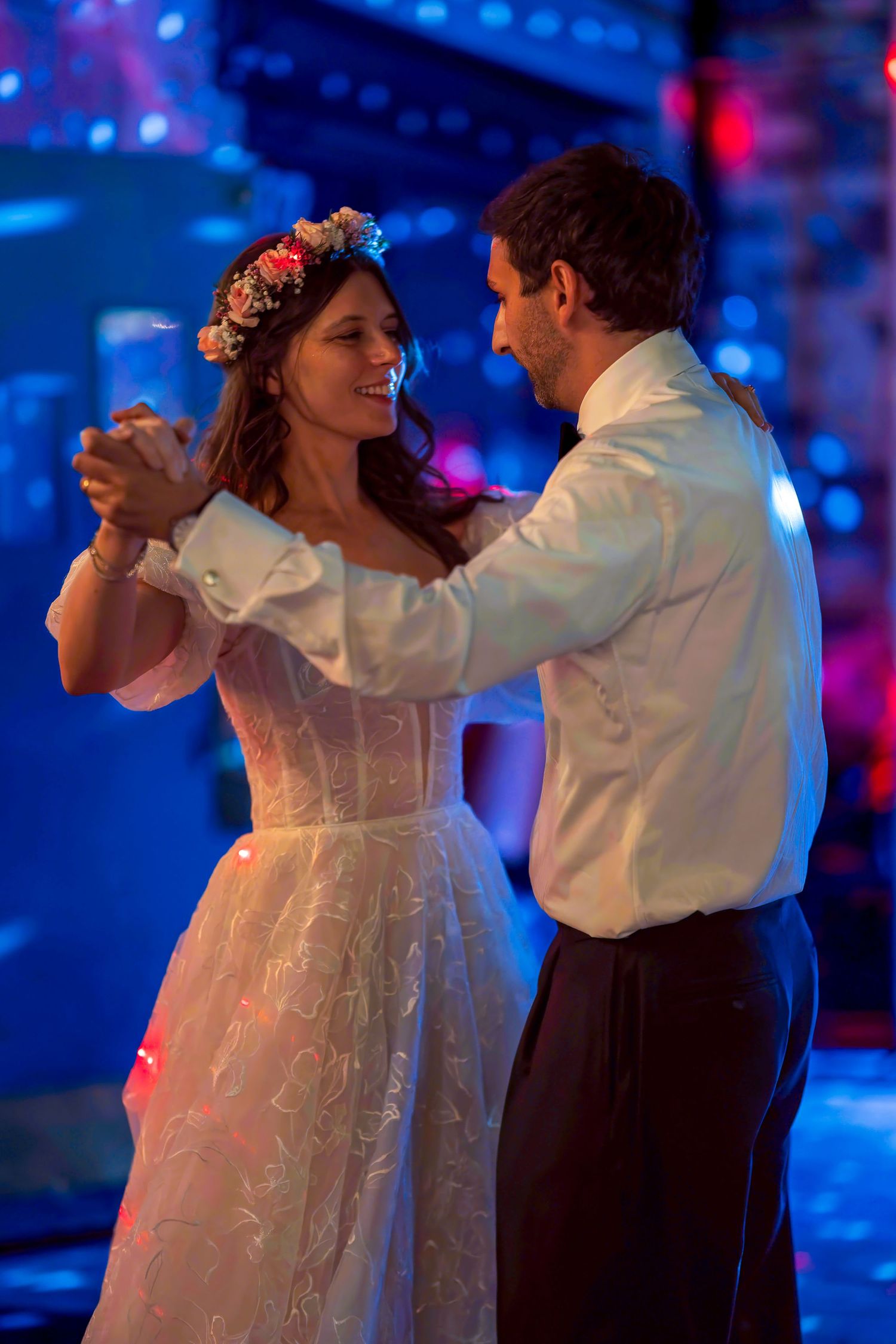 Newlyweds share first dance in blue-lit venue, bride wearing floral crown and off-shoulder wedding dress.