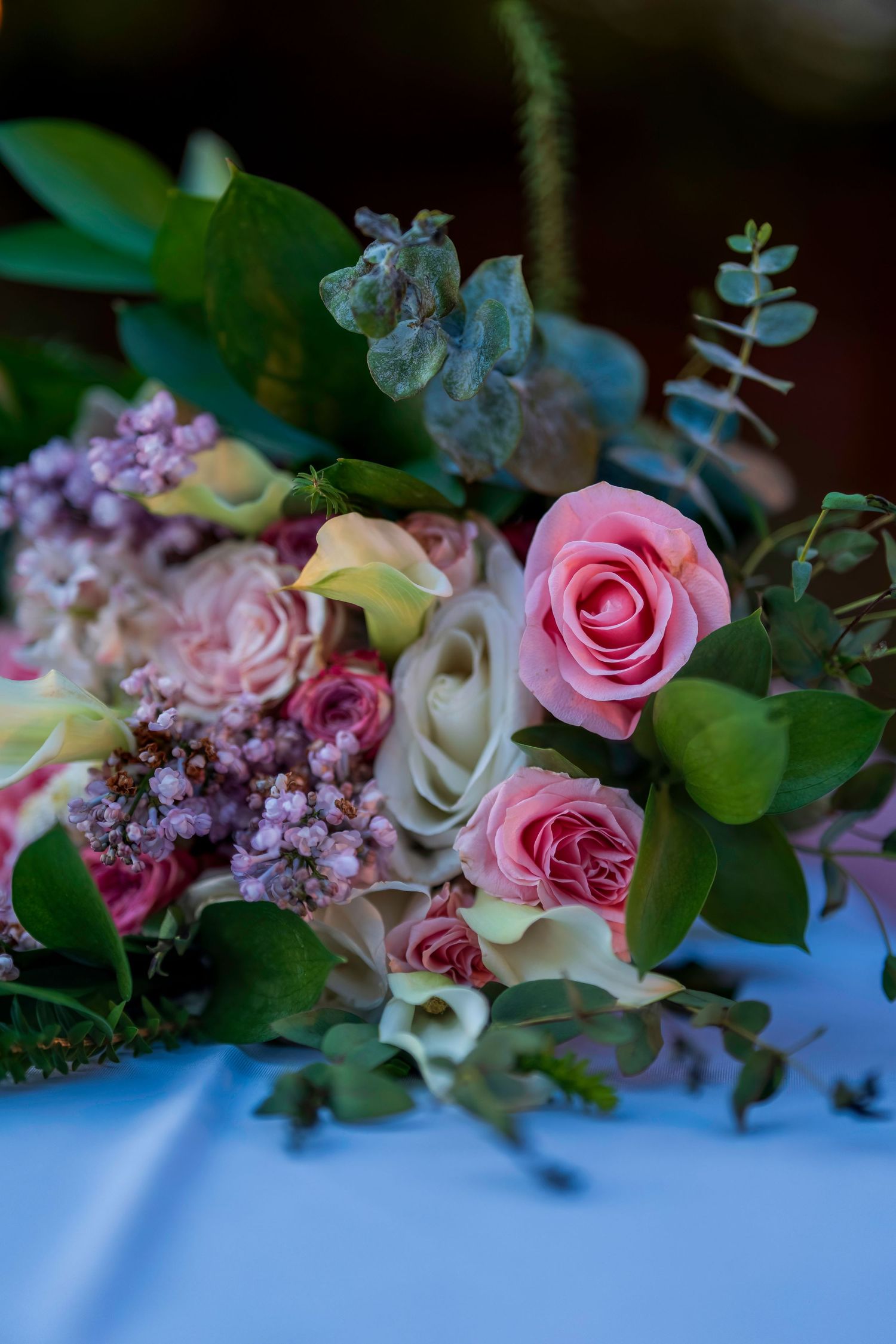 Elegant wedding bouquet with pink roses, white calla lilies, lilac flowers, and eucalyptus greenery on a white table.